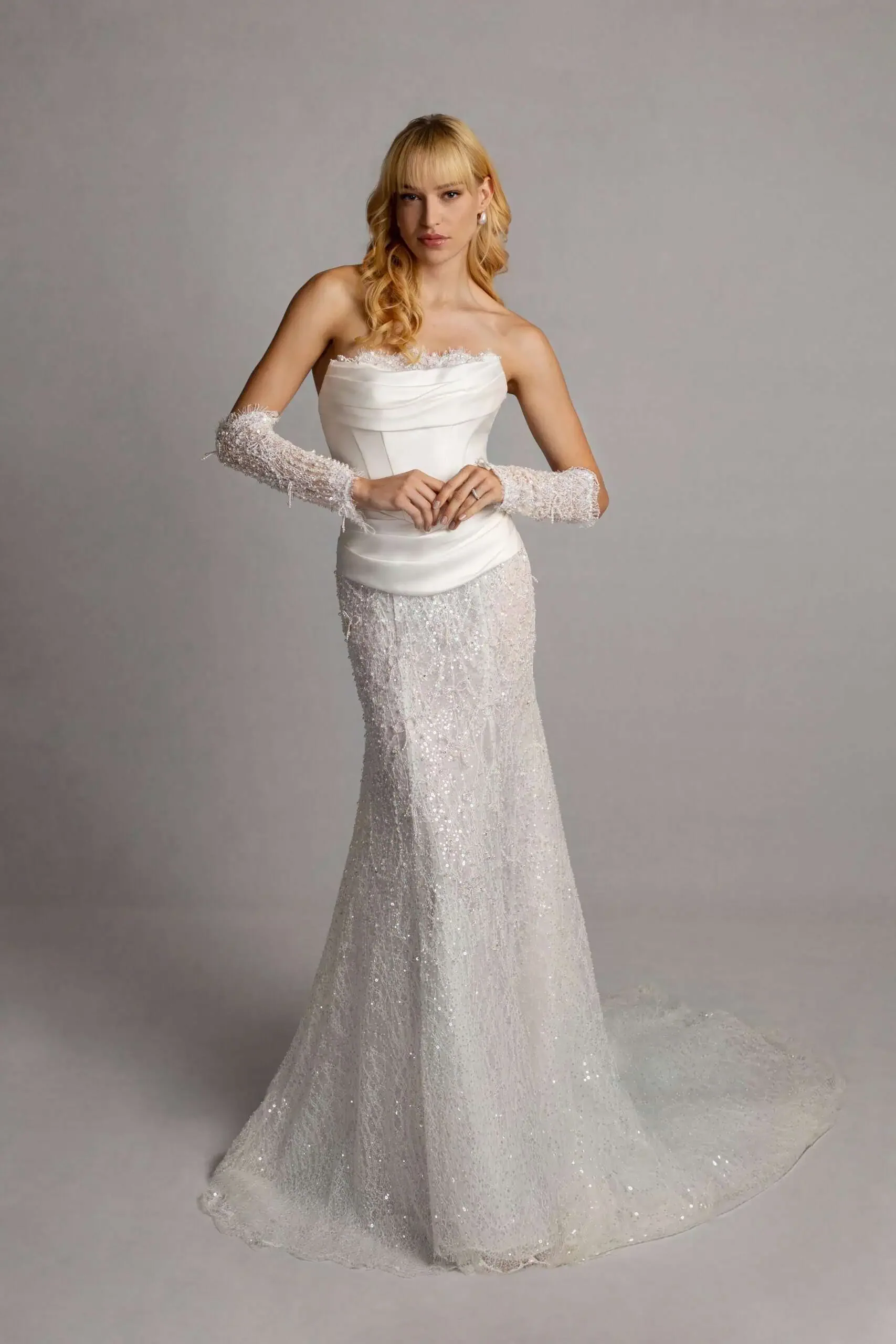 A woman wearing a white wedding gown with lace gloves, standing against a plain gray background.