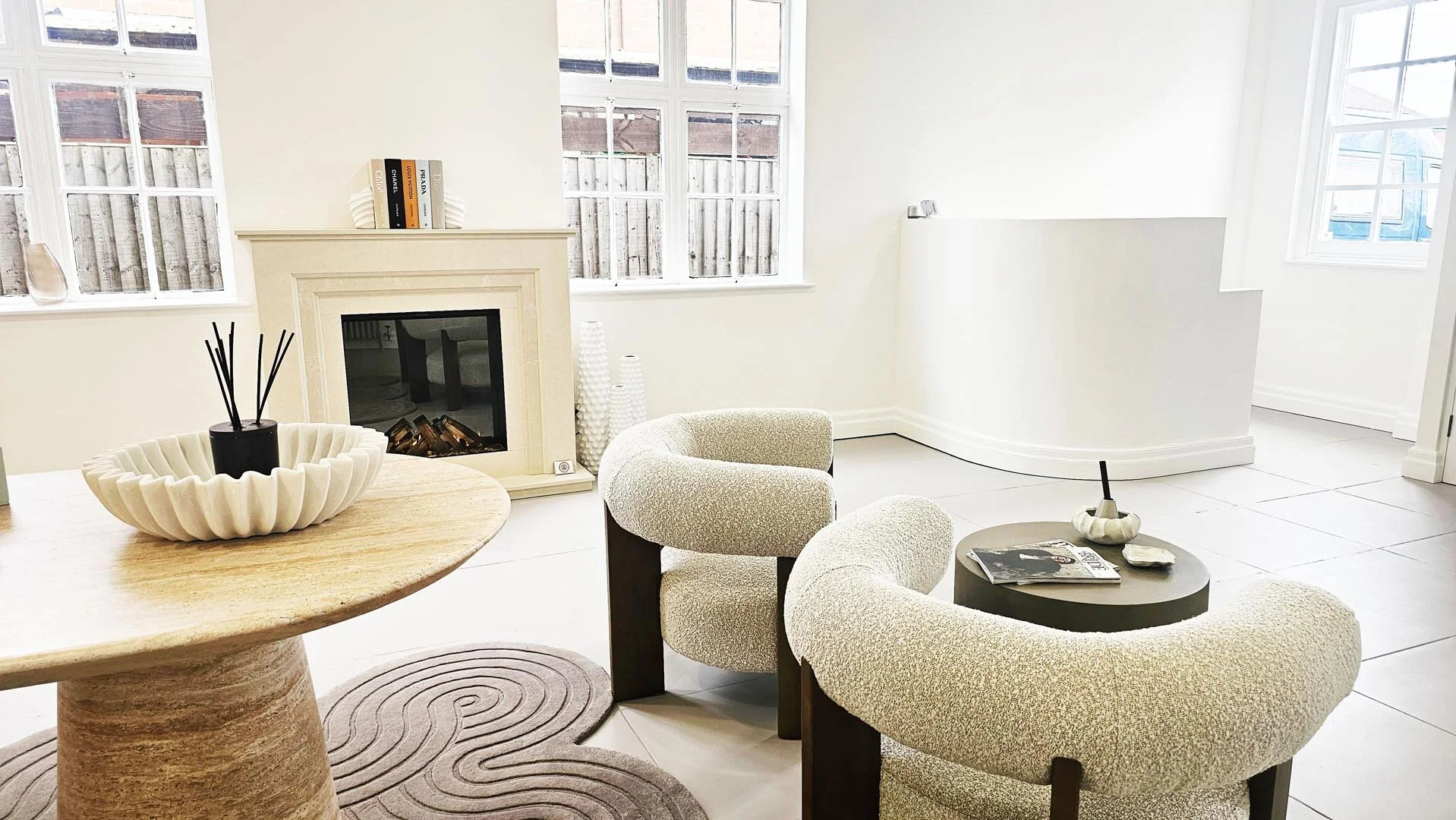 Living room with white walls, fireplace, two beige armchairs, and a round black coffee table with magazines and a decorative item. Large windows with white frames.