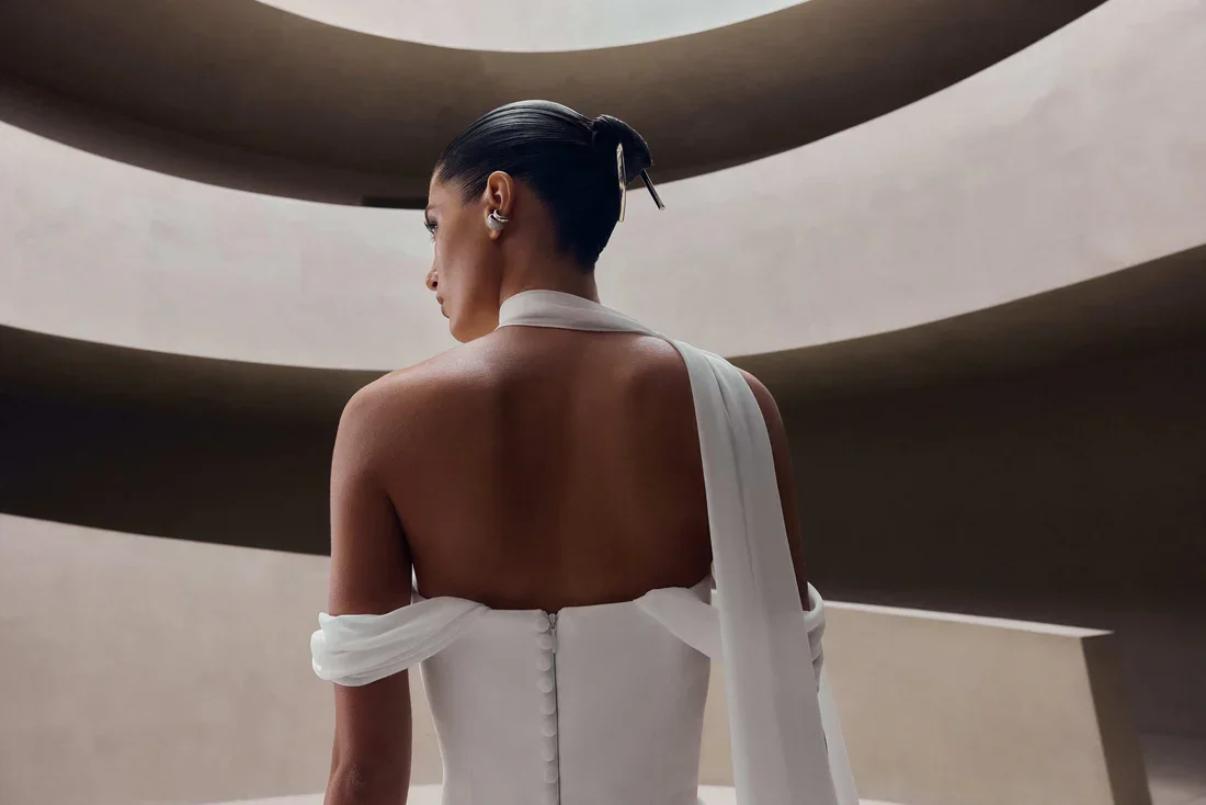A woman with sleek black hair styled in a bun, wearing a white strapless dress with a long, flowing sleeve, standing against a modern architectural background with curved, layered structures.