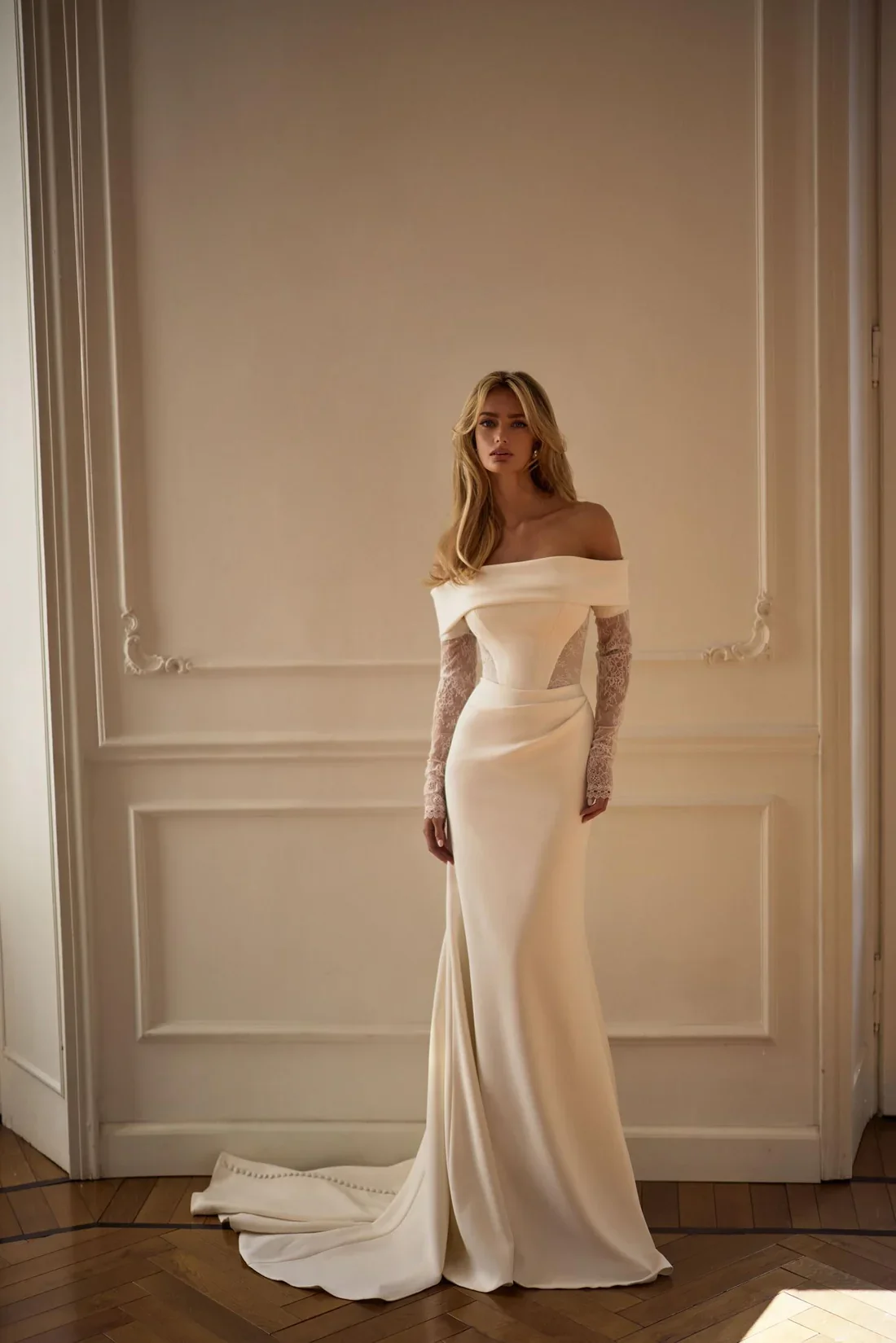 A woman in an off-the-shoulder, long cream-colored gown with lace sleeves, standing in a room with ornate white walls and wooden flooring.