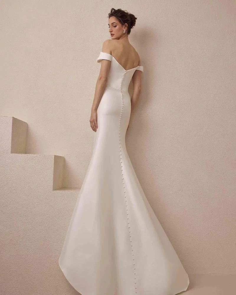 A woman in an elegant off-the-shoulder white wedding gown with buttons down the back, standing against a plain wall.
