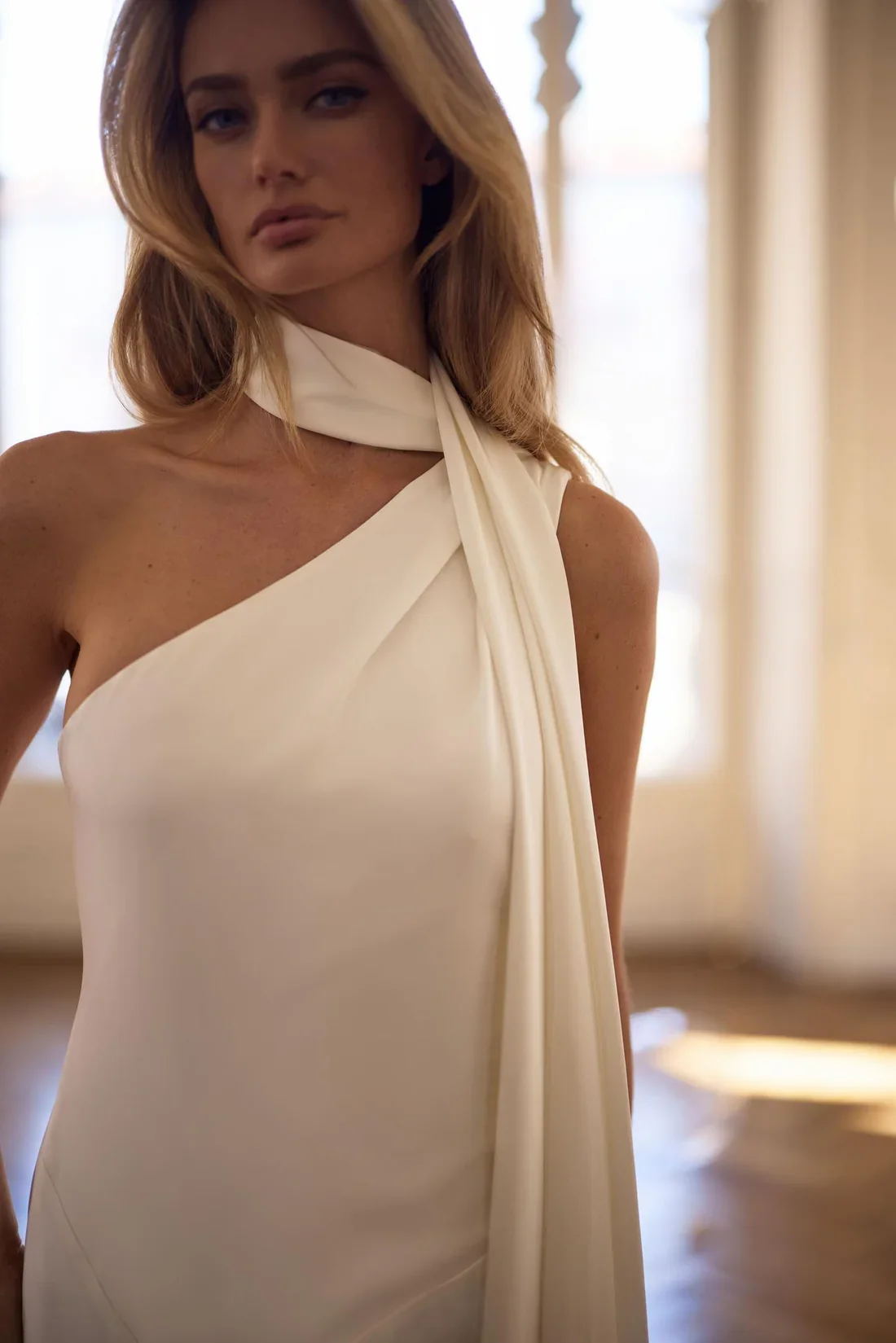 A woman with long, wavy blonde hair standing indoors, wearing a one-shoulder white dress with a draped scarf detail at her neck, in front of a large window with curtains.