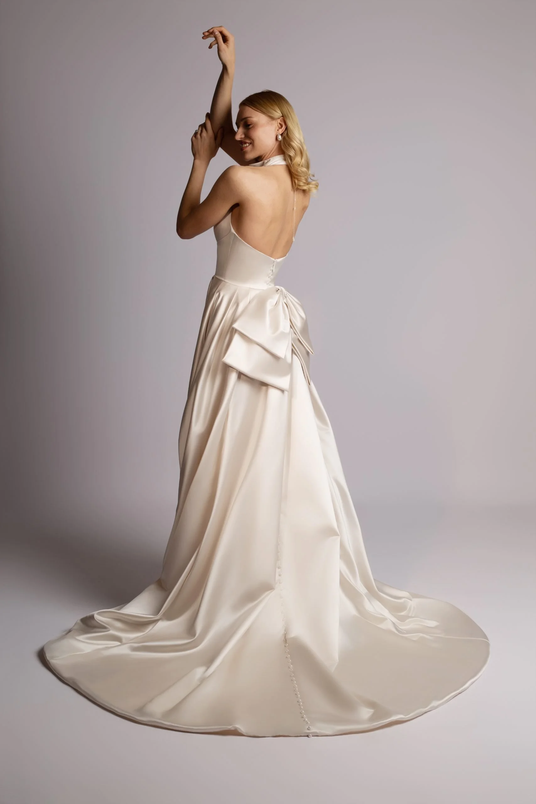 A woman in a backless white satin dress with a large bow on the side, smiling and posing against a plain gray background.
