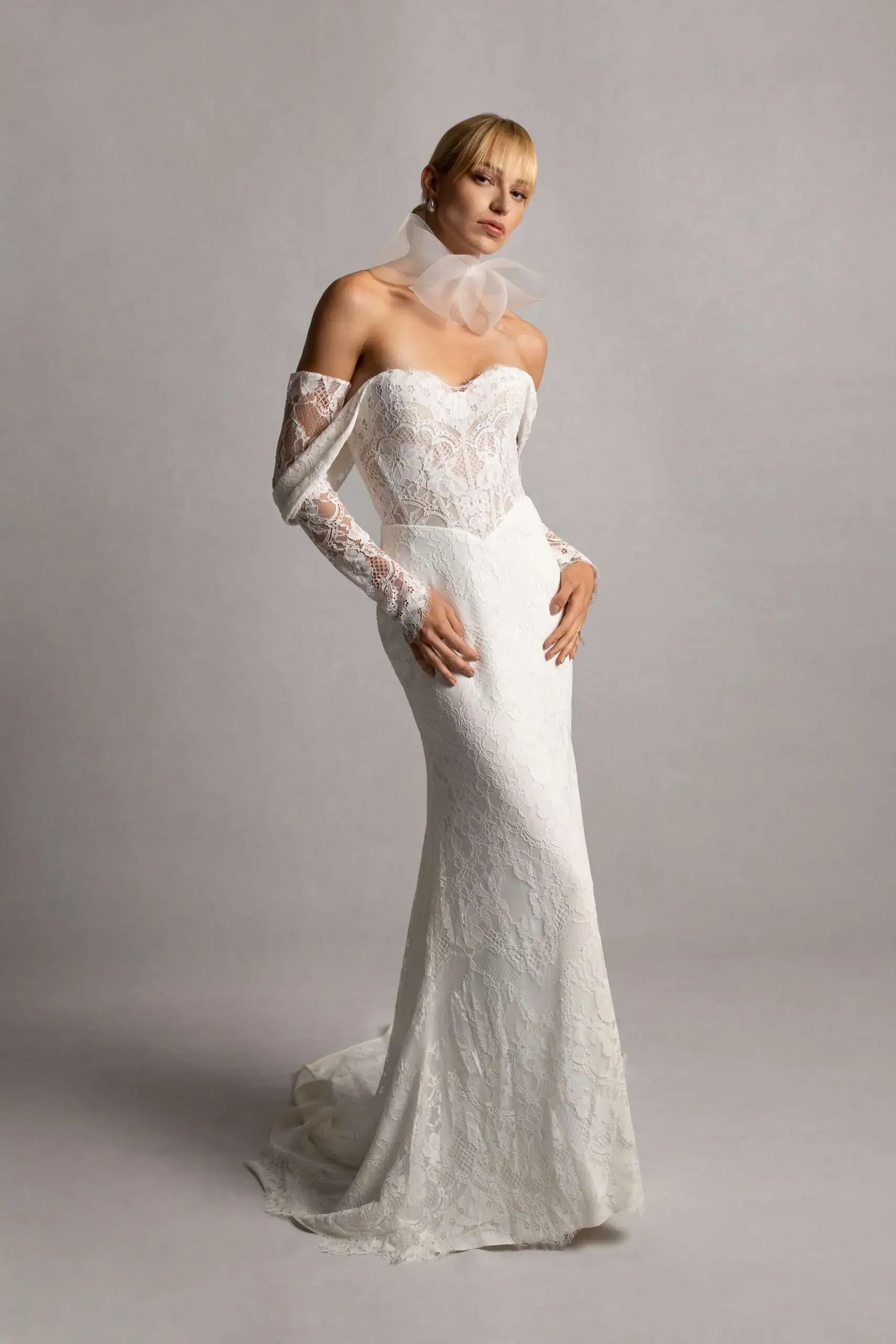 A woman wearing a white lace wedding dress with off-the-shoulder sleeves and a sheer lace overlay, standing against a plain background.