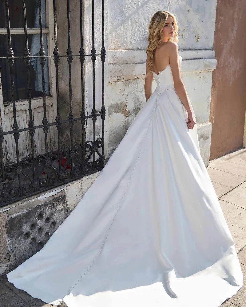 A woman in a white wedding dress with a train, standing outdoors near an aged building with a black wrought iron window grille.
