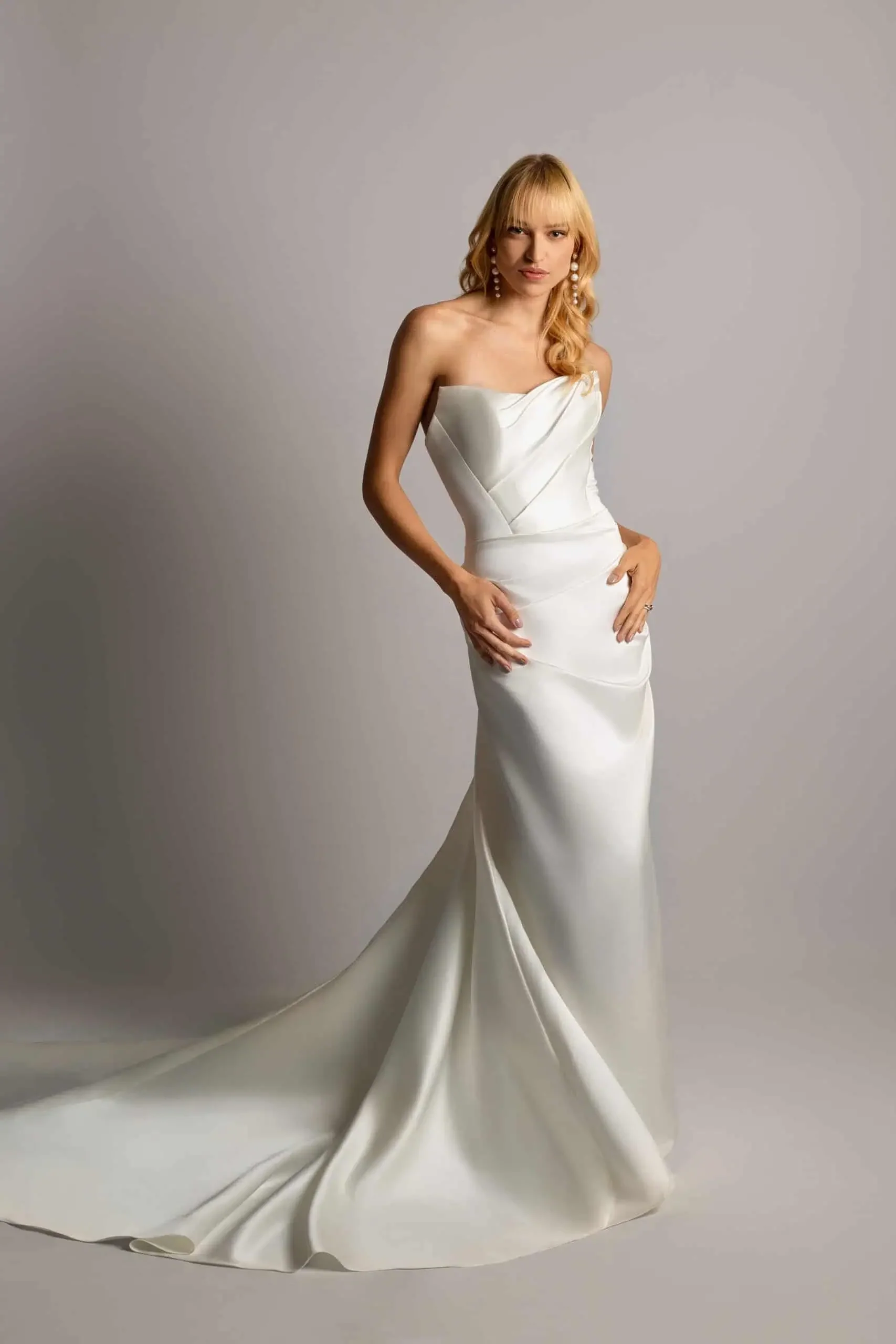 A woman in a strapless white satin wedding dress with a train, standing against a plain light gray background.