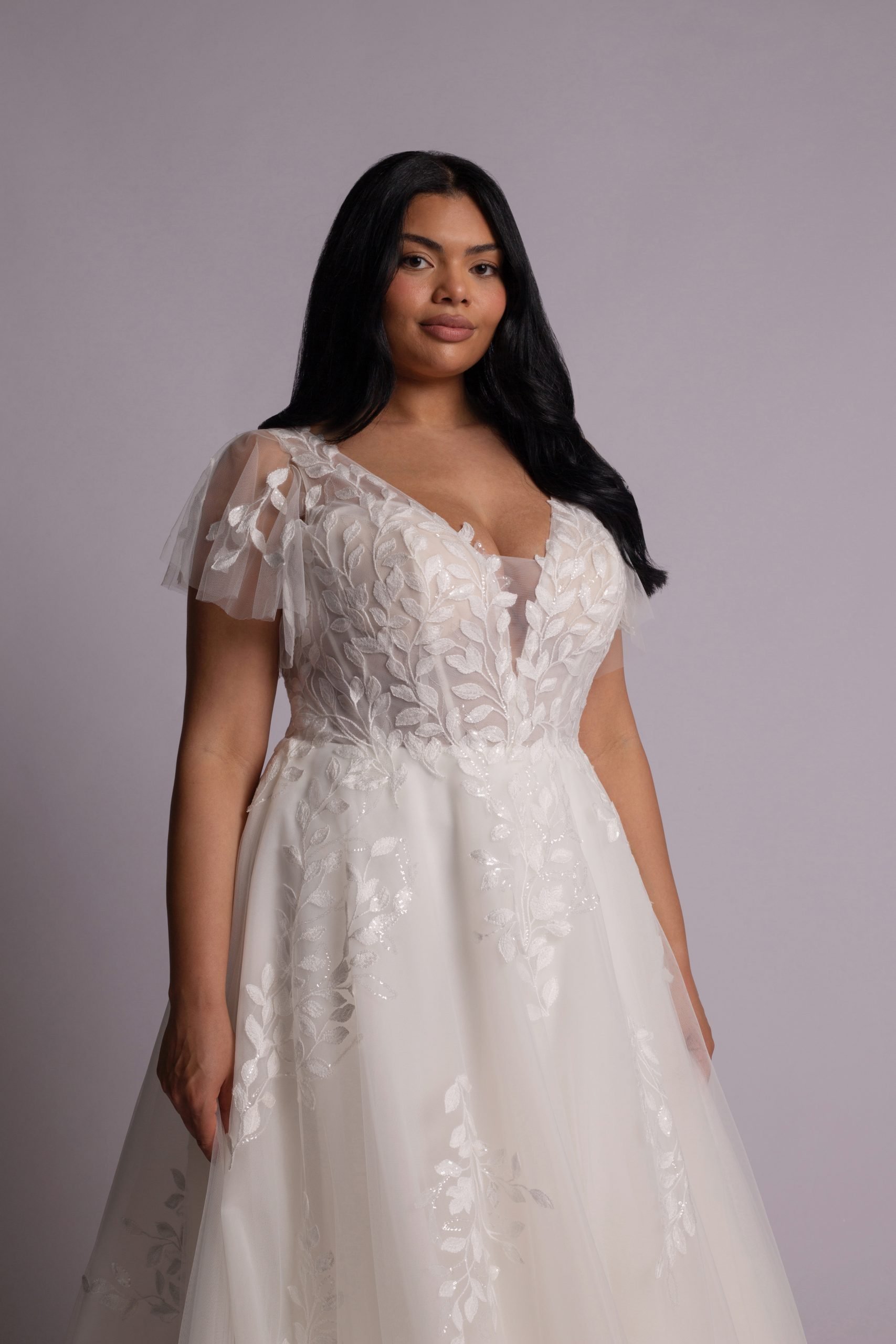 A woman with long black hair wearing an elegant white wedding dress with floral lace details and tulle sleeves.