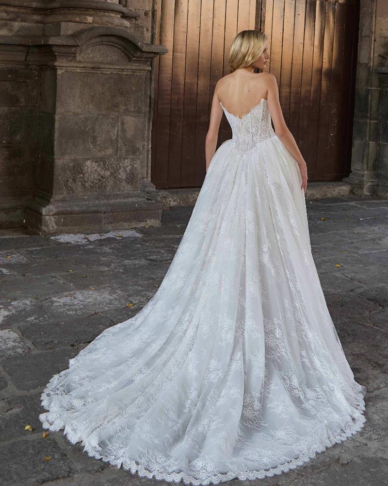 A woman in a white wedding dress with a long train, standing outdoors on a cobblestone surface, facing away from the camera, with a wooden gate and stone wall in the background.