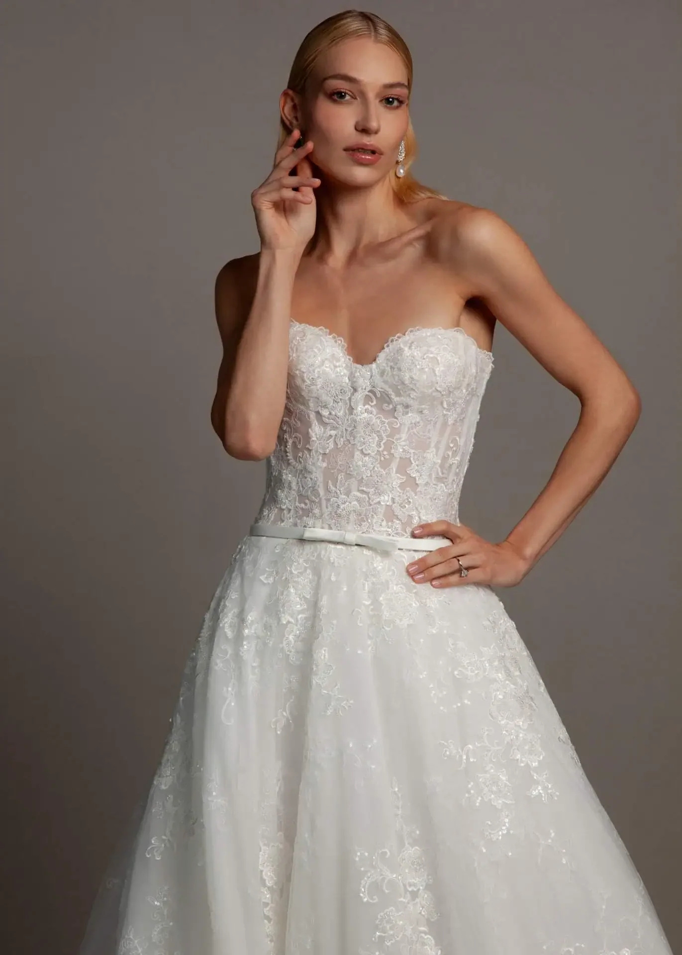 A woman in a white lace wedding dress with a strapless sweetheart neckline, posing against a plain background.