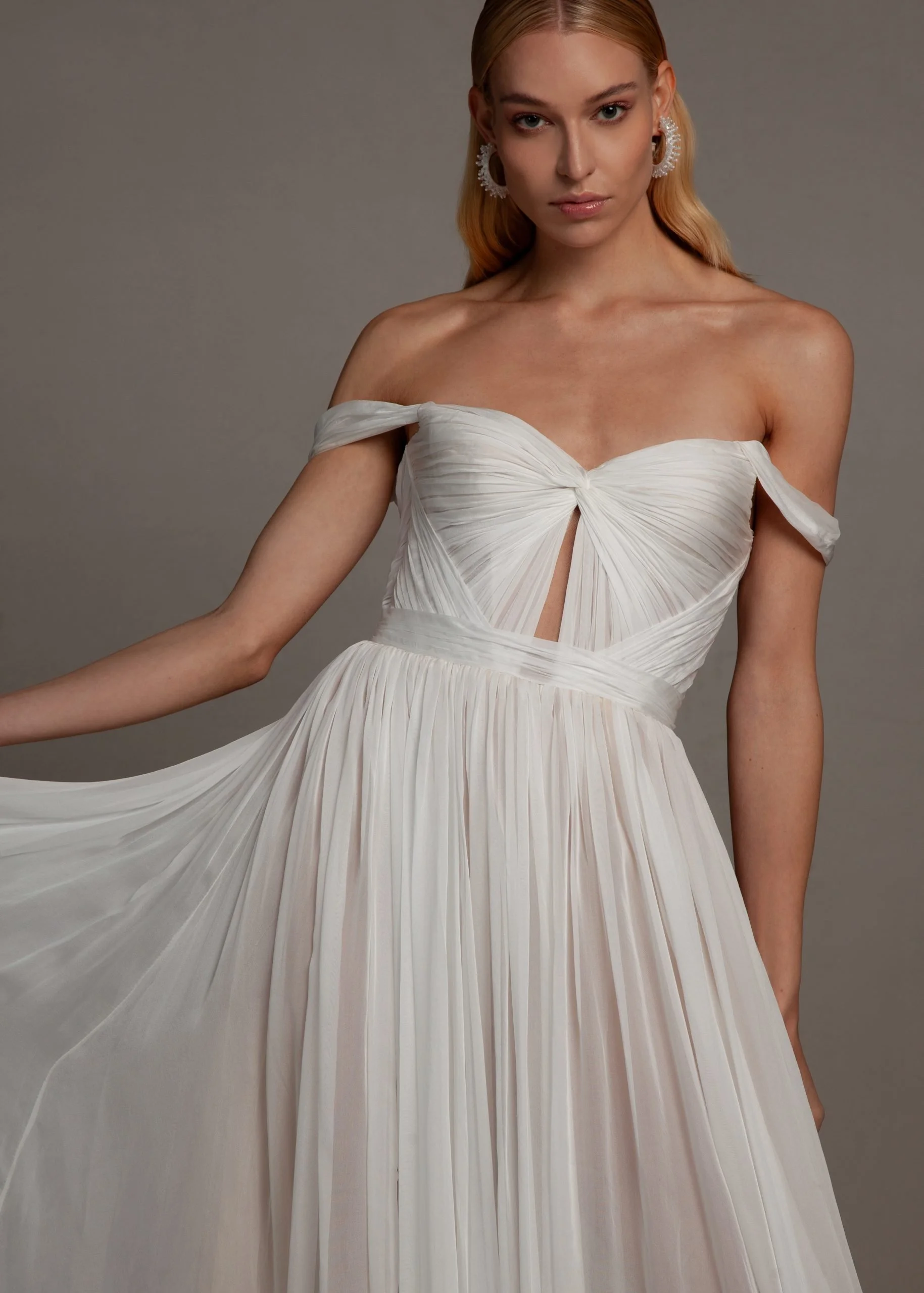 A woman with blonde hair wearing a strapless white pleated dress with off-the-shoulder sleeves and large earrings.