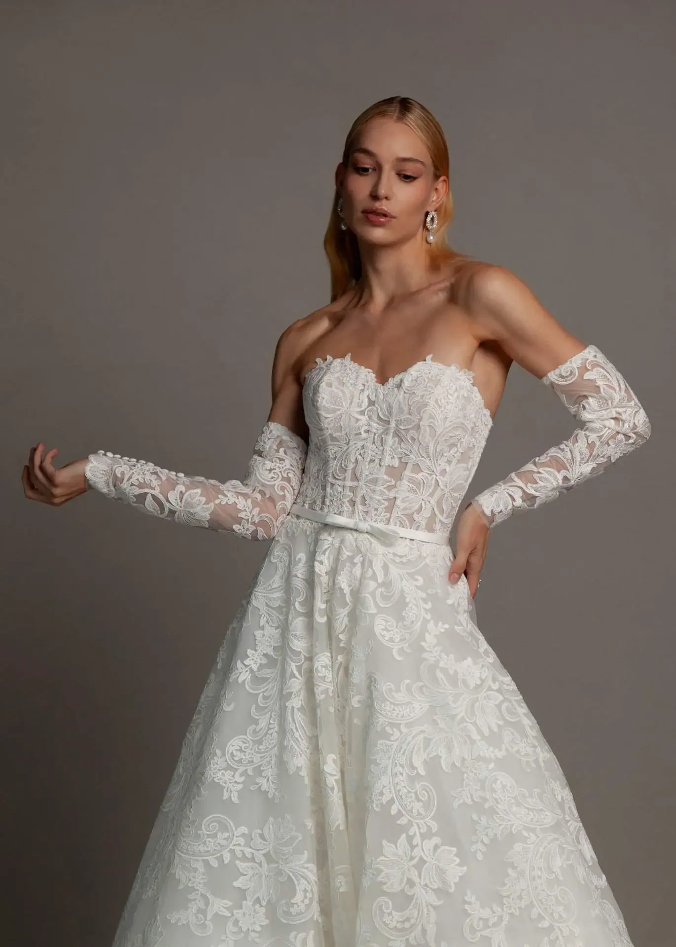 Woman modeling an elegant white lace wedding dress with long lace gloves, earrings, and her hair styled in loose waves against a plain gray background.