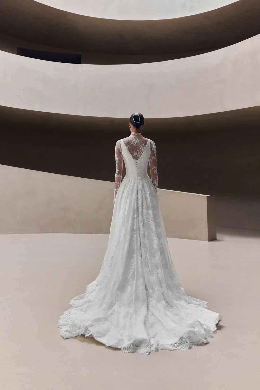 A woman in a long, elegant lace wedding dress standing in a minimalist interior with curved, beige walls, seen from the back.