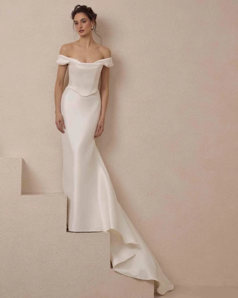 A woman standing on stairs wearing an off-shoulder, white satin wedding dress with a fitted silhouette and train.