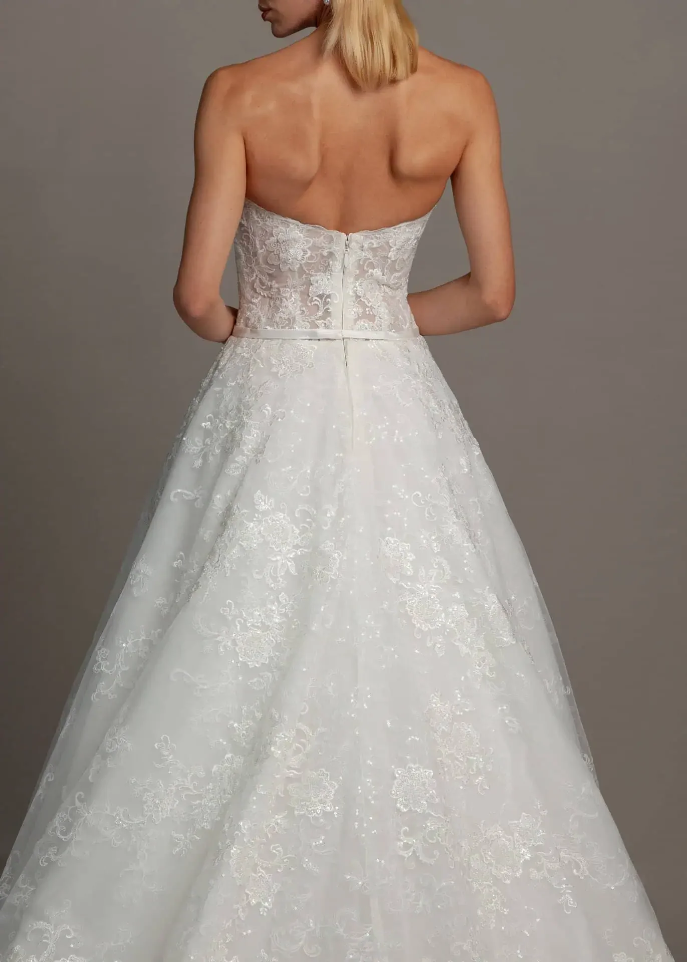 Woman in a strapless white wedding gown with lace detailing, standing against a plain gray background, viewed from the back.