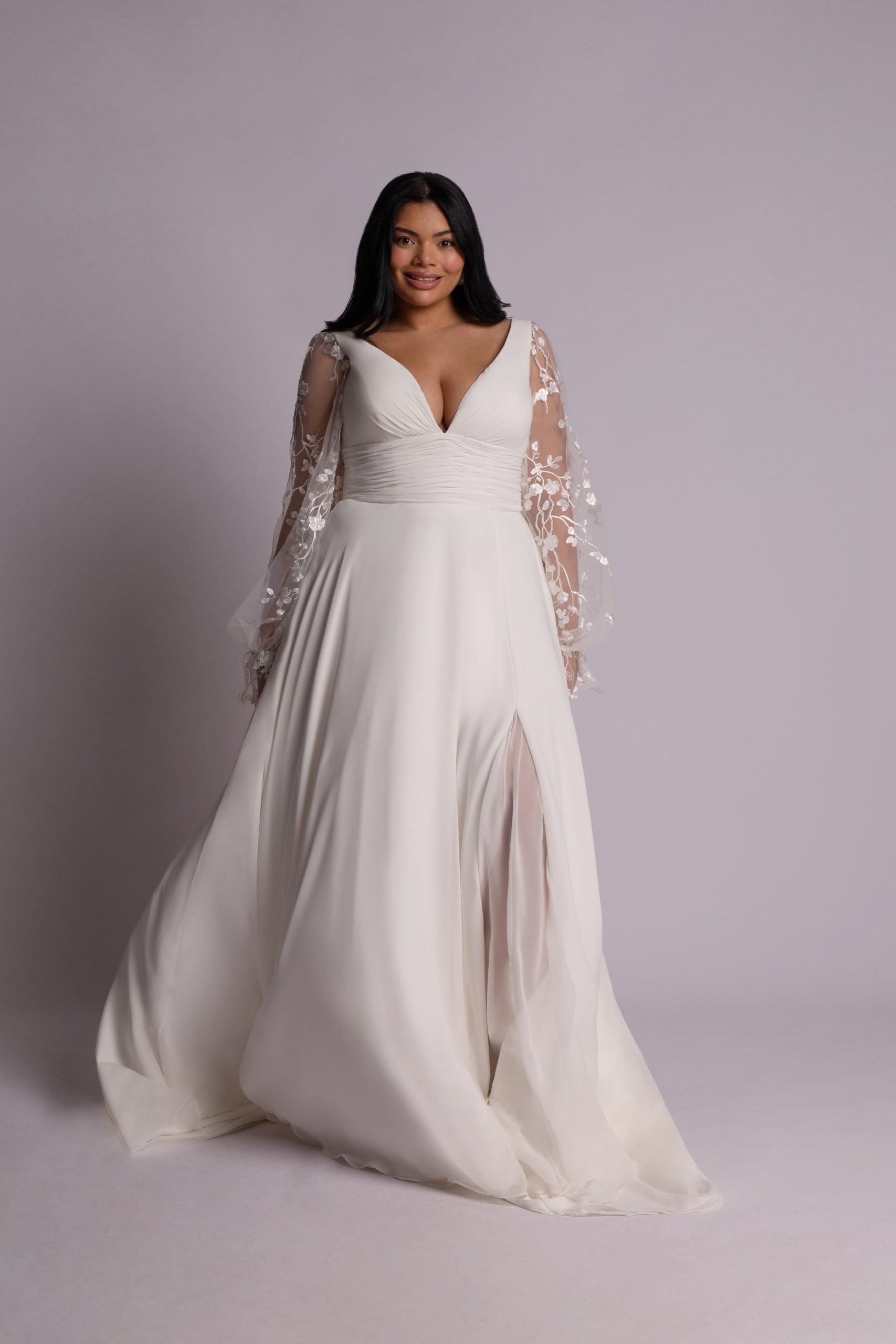 A woman wearing a white wedding dress with lace sleeves standing against a plain background.