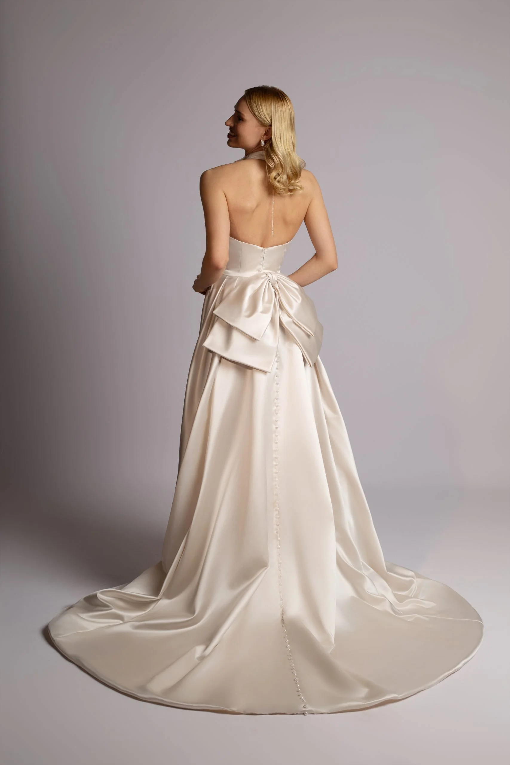 A woman wearing a strapless, satin wedding gown with a large bow at the back, standing on a plain gray background.