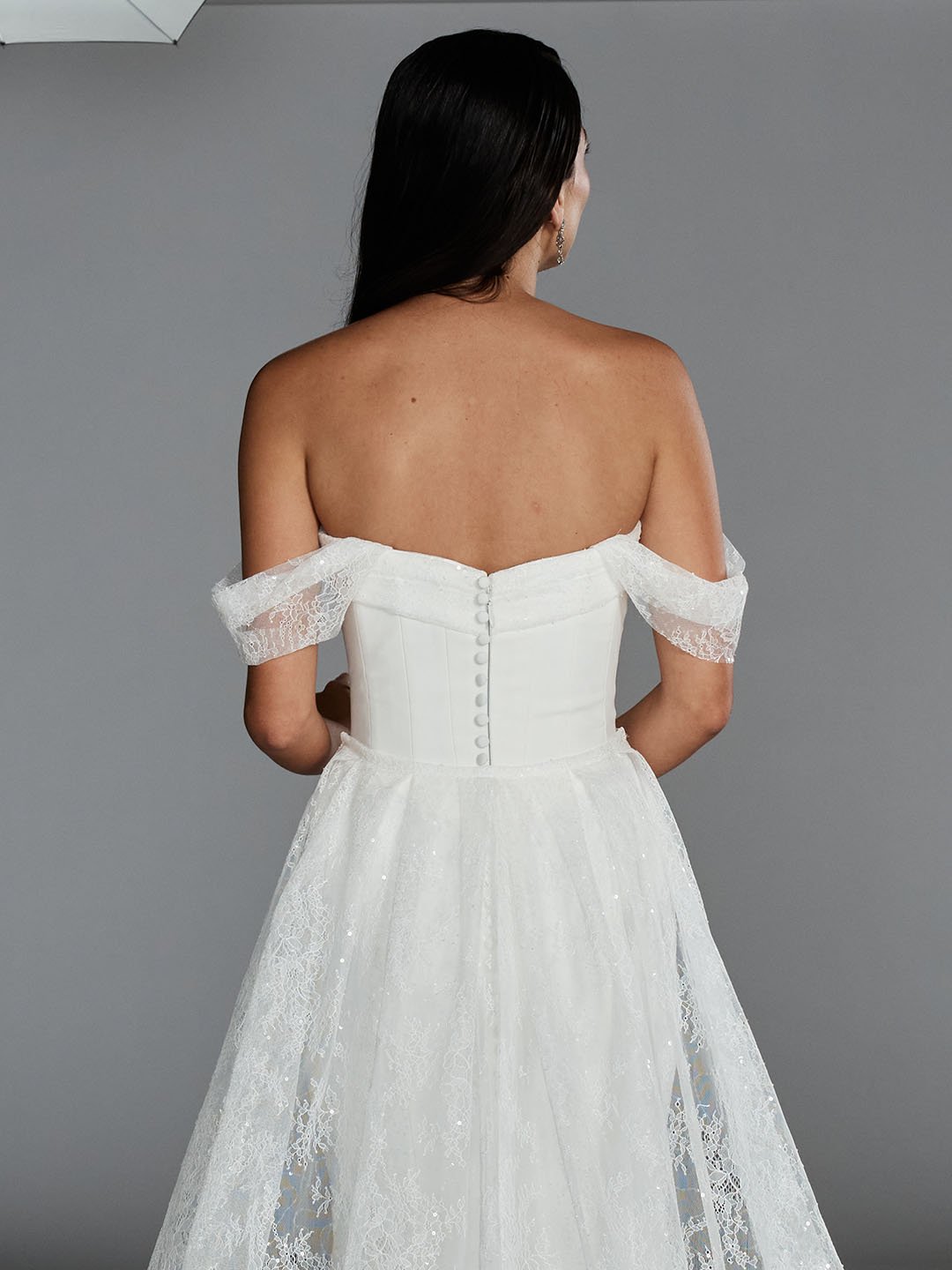 Back view of a woman in a white strapless wedding dress with lace off-the-shoulder sleeves and buttons running down the back.