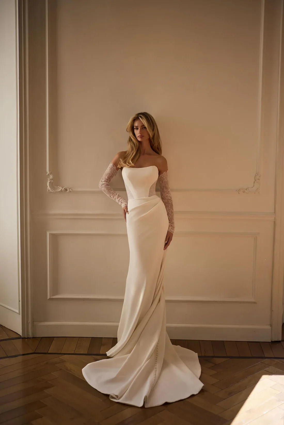 A woman in a white wedding gown with lace long sleeves stands in an elegant room with beige walls and hardwood floors.