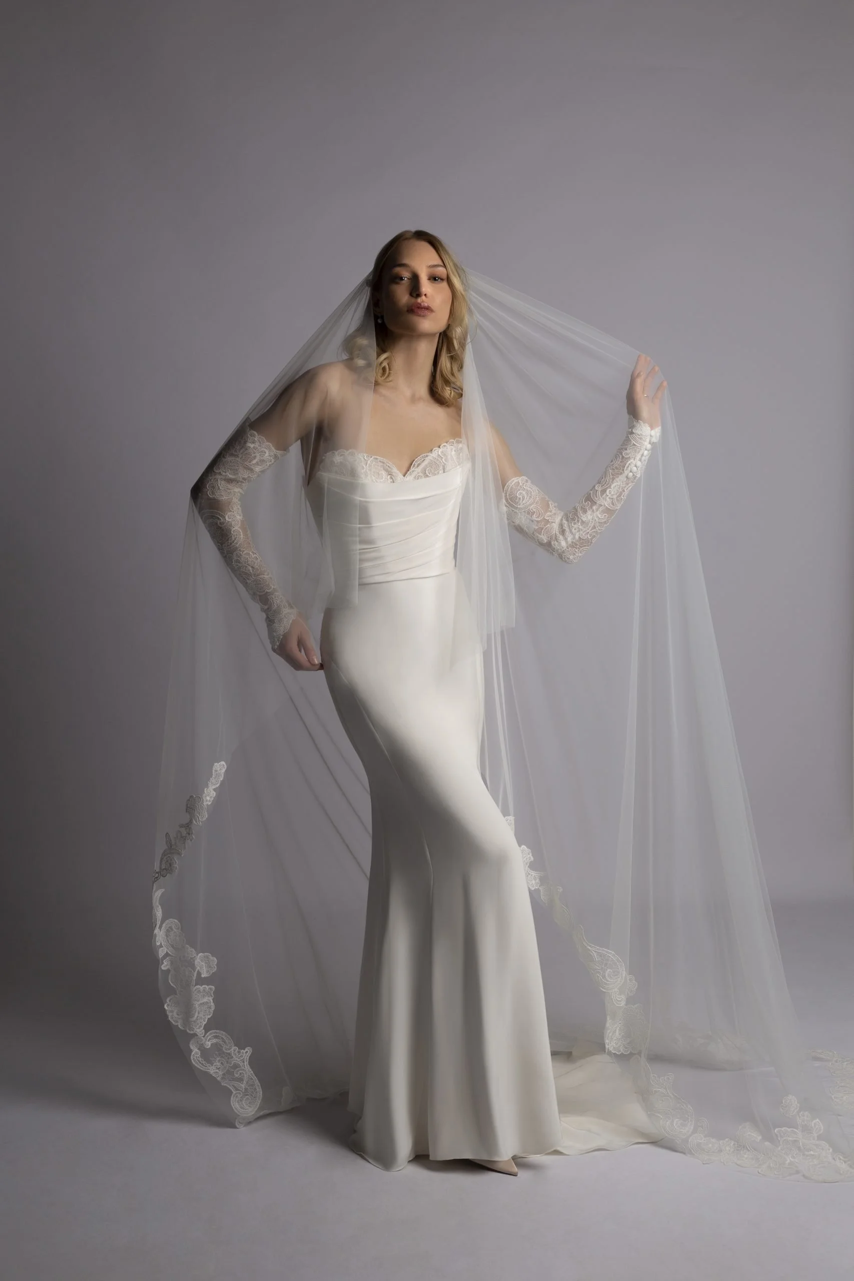 A woman in a strapless wedding dress and lace gloves, posing with a veil against a plain gray background.