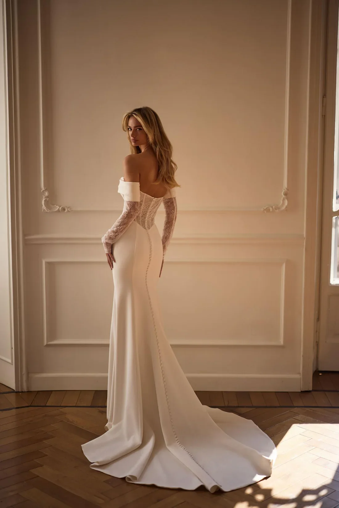 A woman in a wedding dress with lace sleeves and a long train, standing in a softly lit room with beige walls and wooden flooring.
