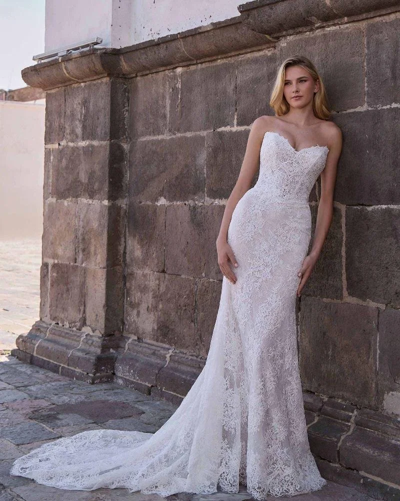 A woman in a strapless white lace wedding dress standing against a stone wall outside, with a small train extending on the ground.