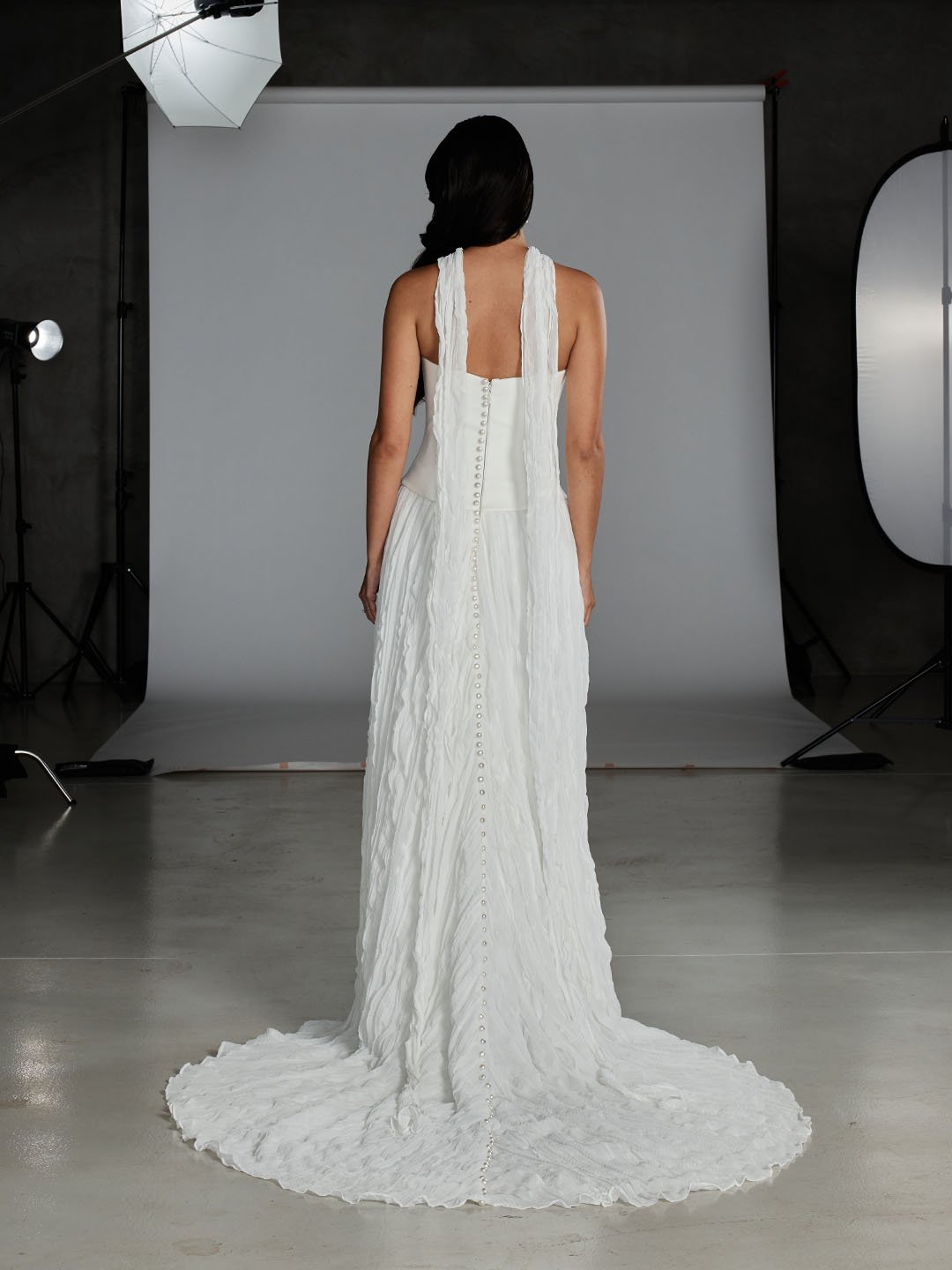 Back view of a woman wearing a long, white, textured dress with buttons down the back in a photography studio.