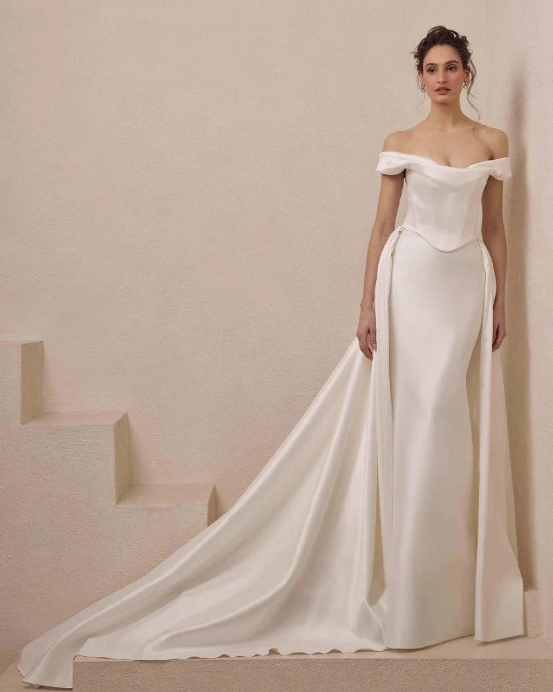 A woman in a white off-shoulder wedding gown with a long train, standing against a beige wall.
