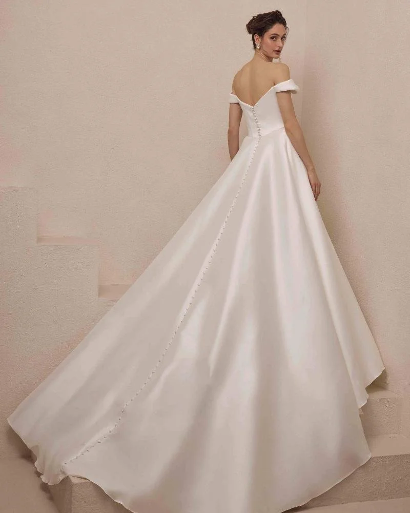A woman in a white, off-the-shoulder wedding gown with a fitted bodice and button detailing down the back, standing against a plain, light-colored wall.