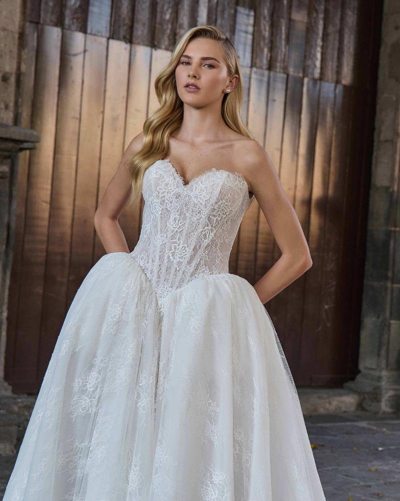 A woman in a strapless white lace wedding dress with a fitted bodice and full skirt, standing outdoors against a wooden fence.