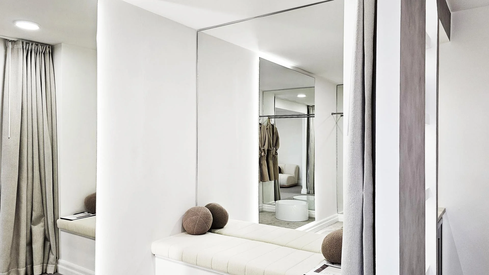 Modern interior space featuring white walls, a cushioned window seat with round decorative pillows, a large mirror, and beige curtains.