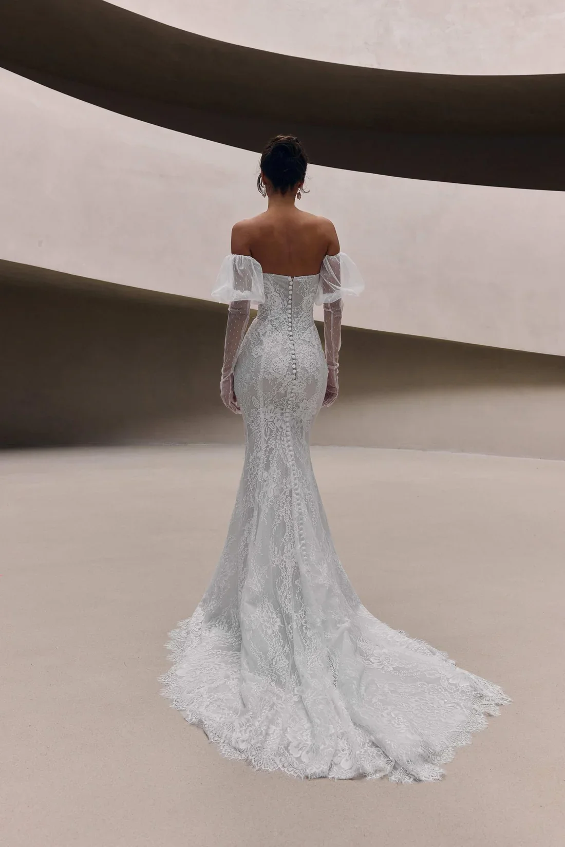 A woman wearing a fitted, off-the-shoulder lace wedding gown with a train, standing with her back to the camera in a modern, minimalistic interior.