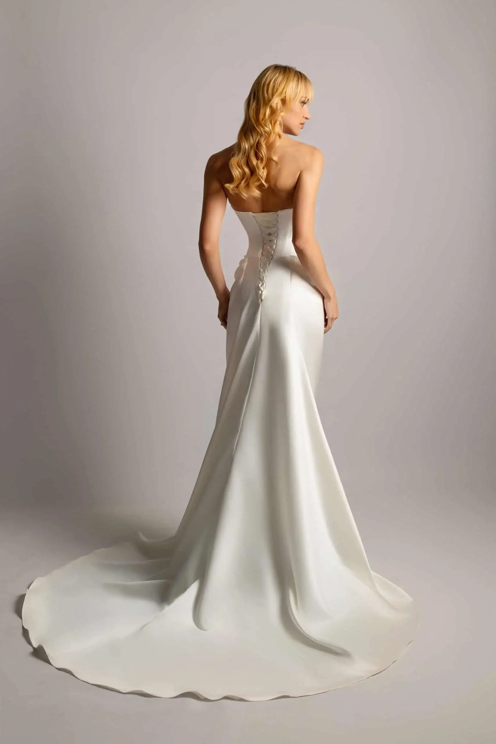 A woman with long, wavy blonde hair wearing a strapless, satin, white wedding gown standing against a plain, light-colored background.
