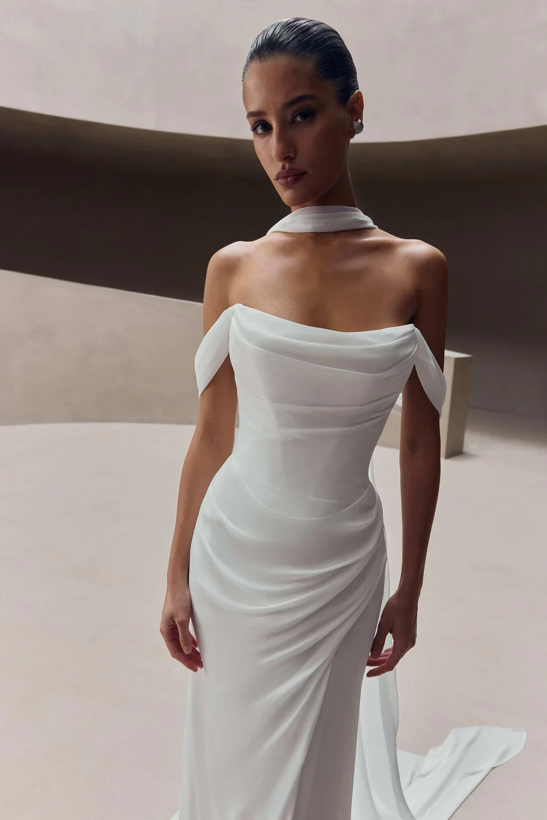 A woman wearing an elegant white off-shoulder dress with draped fabric, standing in a modern, minimalist interior setting.