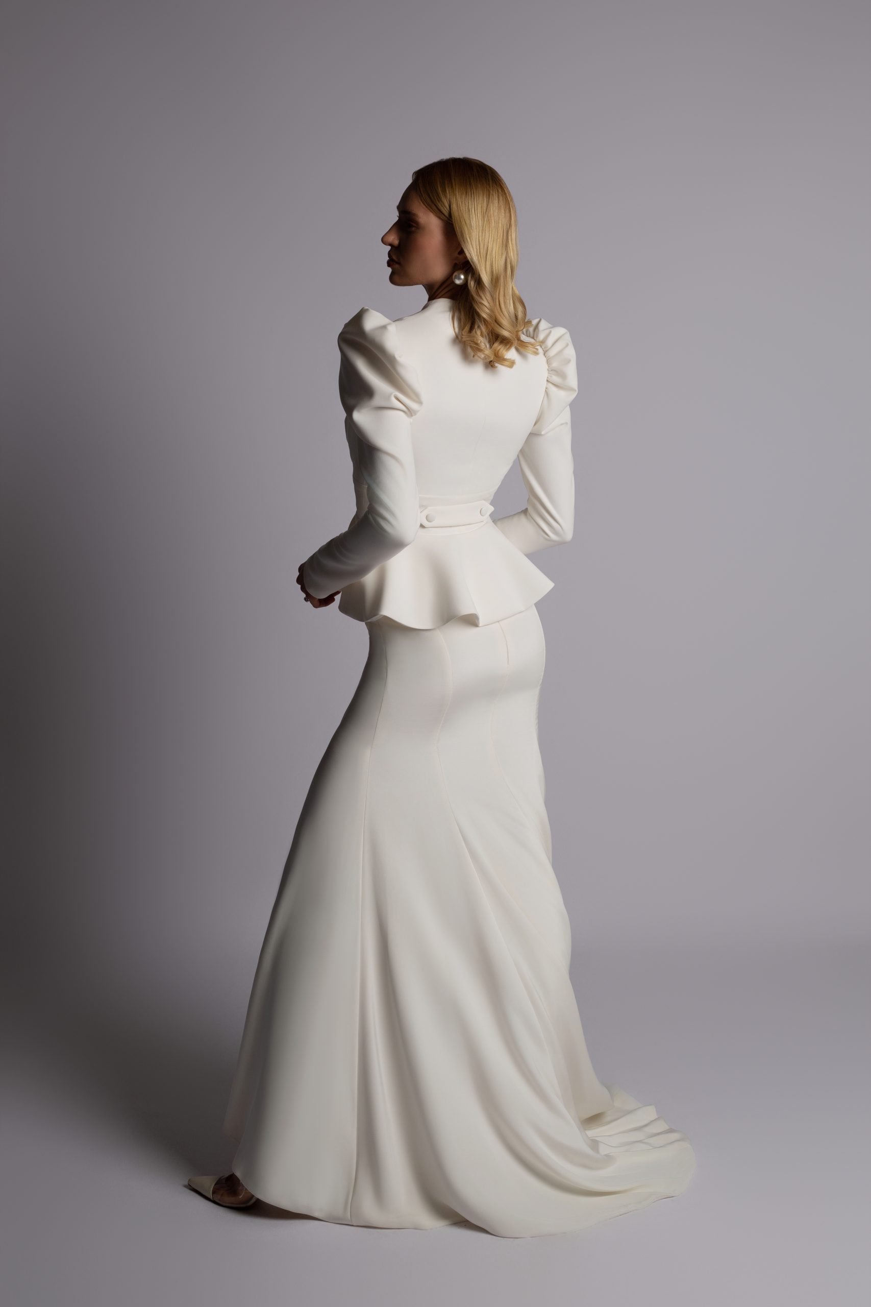 A woman with blonde hair in a white tailored standing dress with puffed shoulders, standing against a plain gray background.