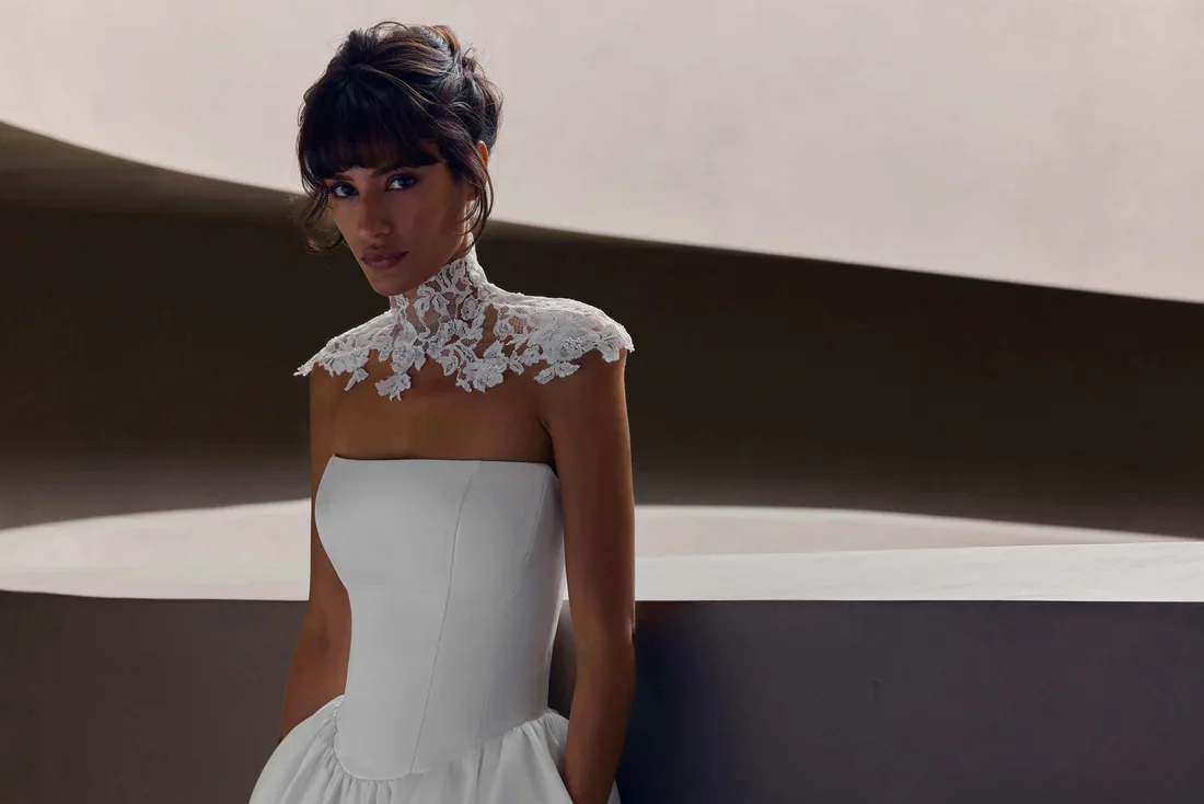 Woman in a strapless white dress with lace flower detail on the neckline, standing against a minimalist background.