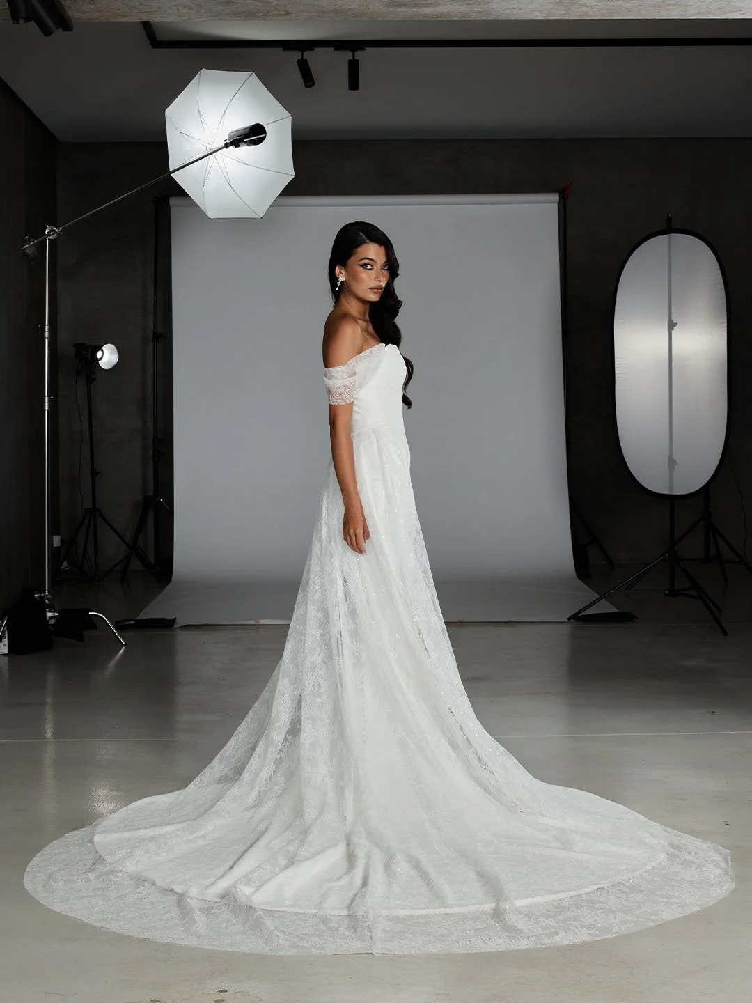 A woman in a white lace off-shoulder wedding dress standing in a photography studio with a gray backdrop, studio lights, and reflective umbrella, looking to the side.
