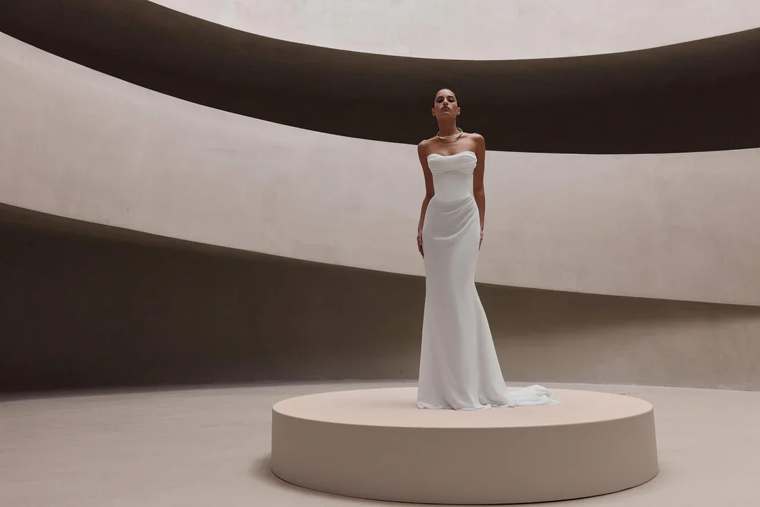 A woman wearing a white strapless wedding gown standing on a round platform in a modern, minimalistic architectural setting with curving lines.