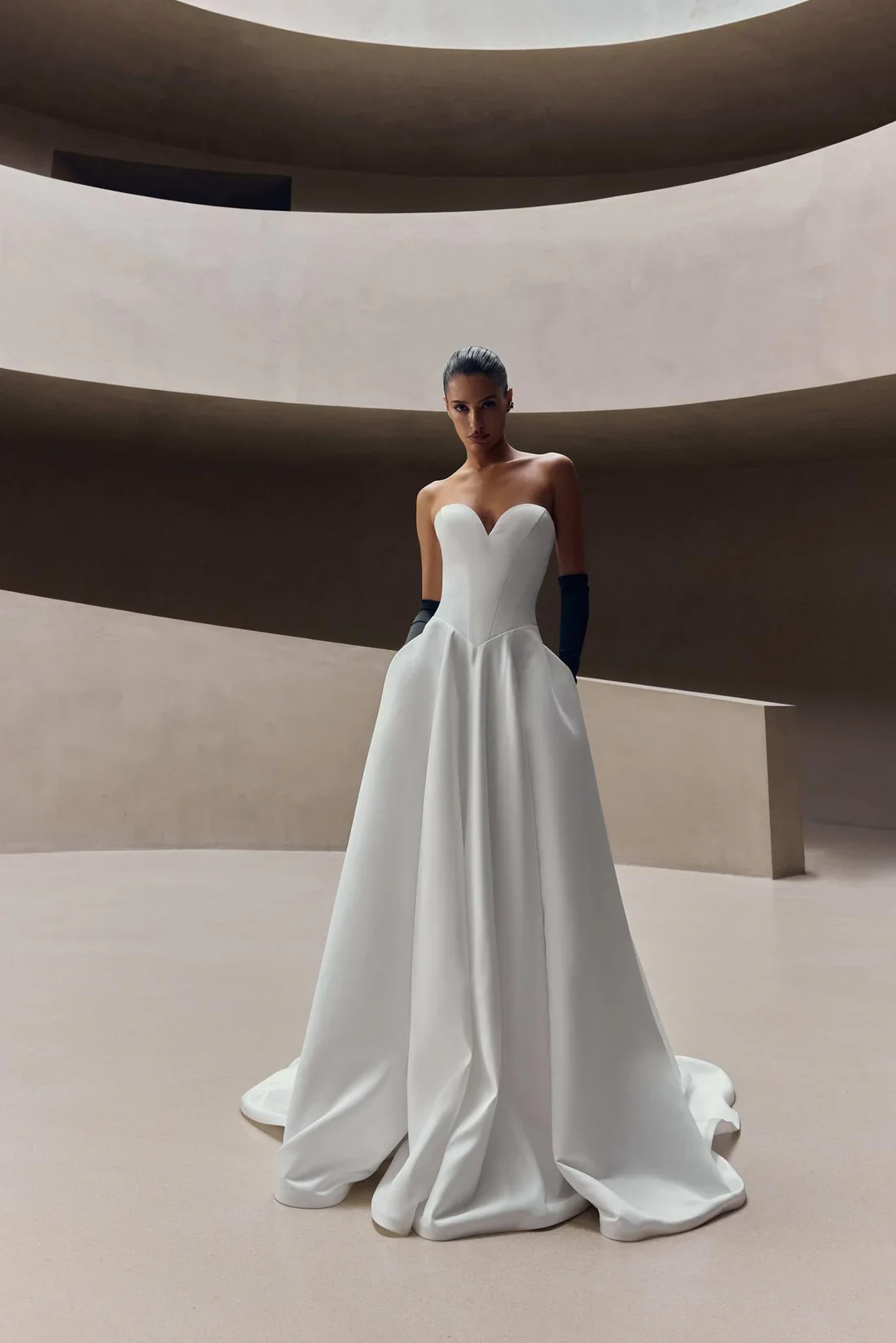 A woman in a strapless white wedding gown standing in a modern, minimalist interior with curved beige walls.