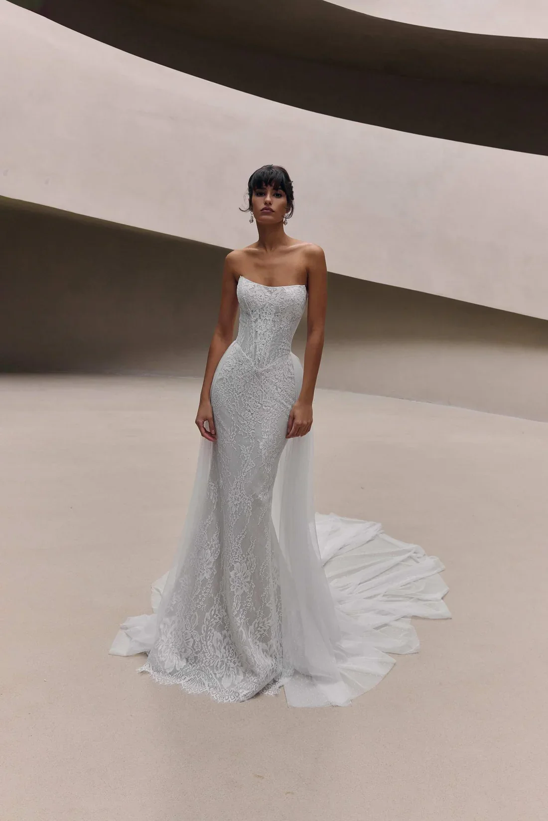 Woman in a white wedding gown with lace detail, standing on a neutral-colored floor inside a modern architectural space with curved, smooth concrete walls.