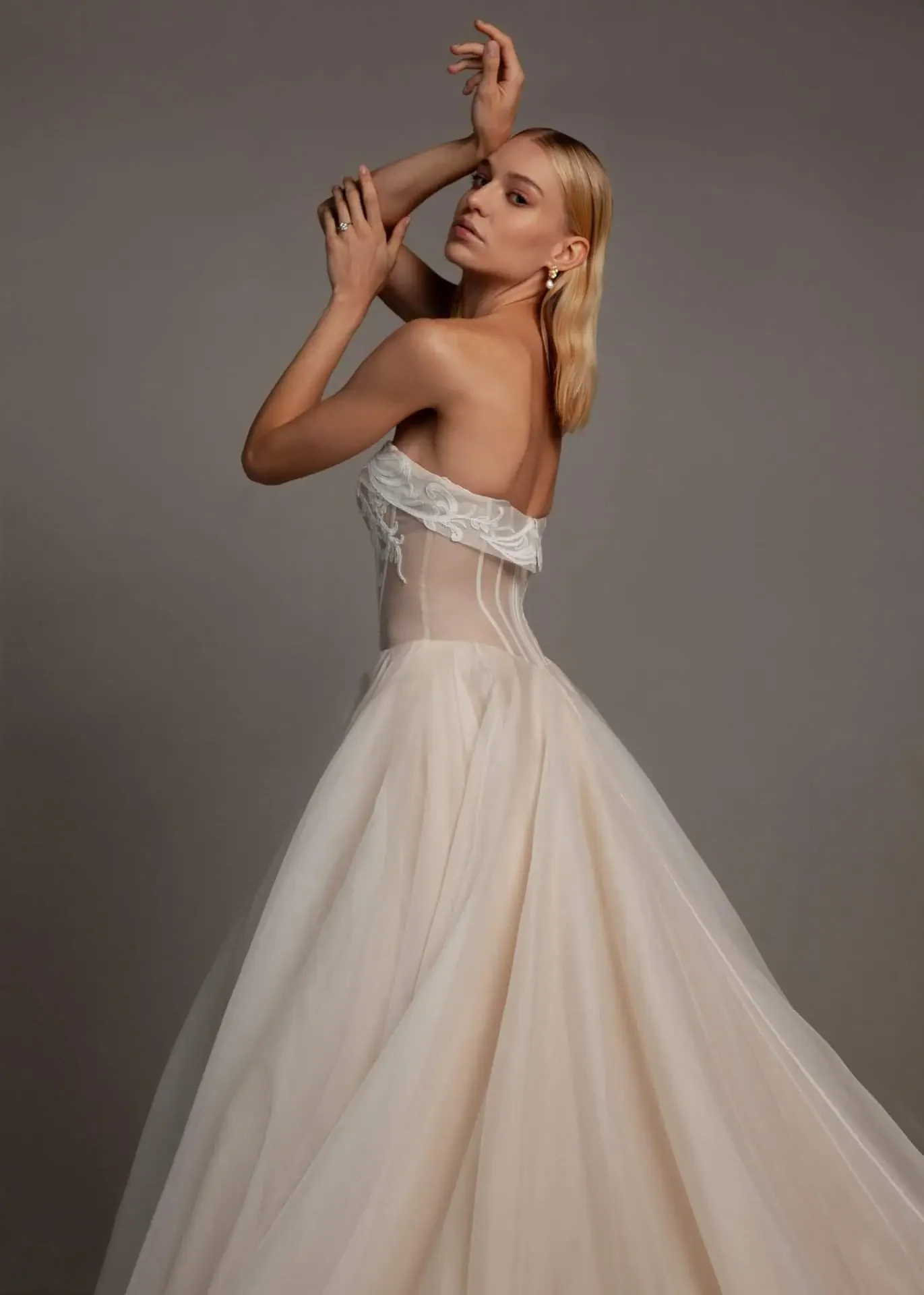 A woman wearing a strapless, cream-colored gown with lace details on the top and a full skirt, posed against a plain background.