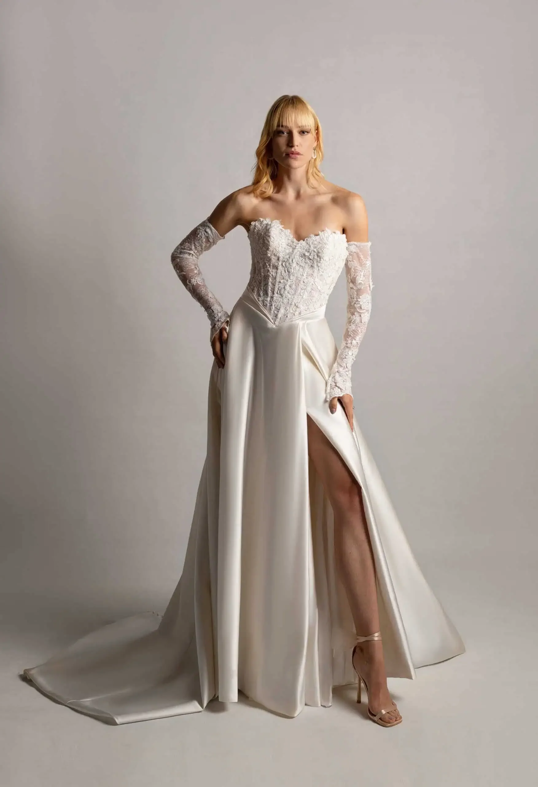 A woman in a white wedding dress with lace sleeves and a high slit, standing against a plain backdrop.