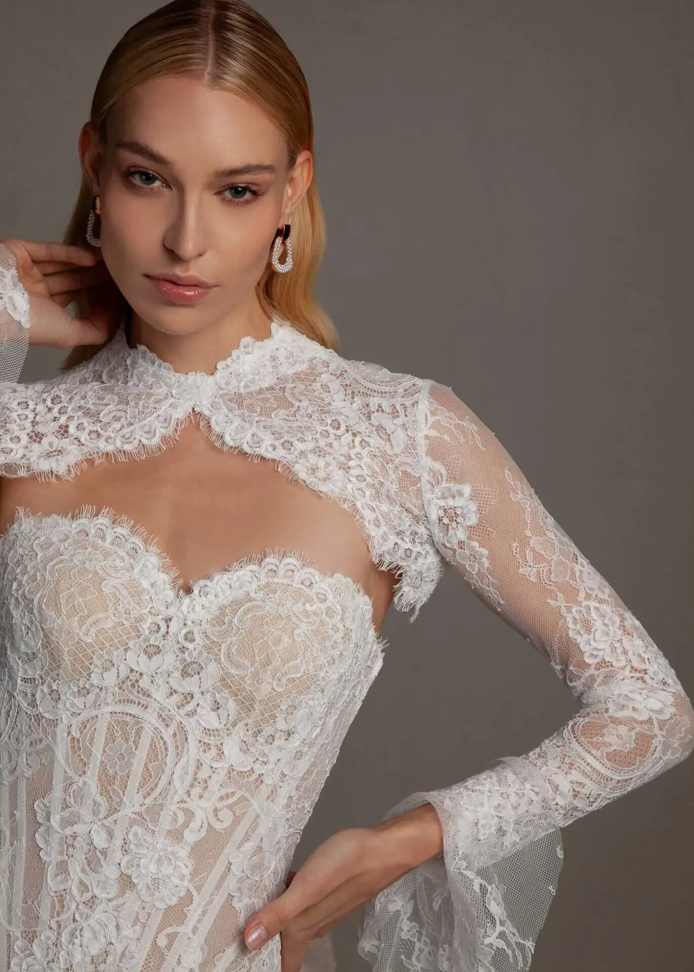 A woman wearing an elegant white lace dress with intricate embroidery and long sleeves, posing against a plain background.