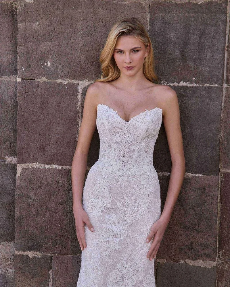 A young woman with blonde hair and light skin is wearing a strapless white lace wedding dress and standing against a stone wall.