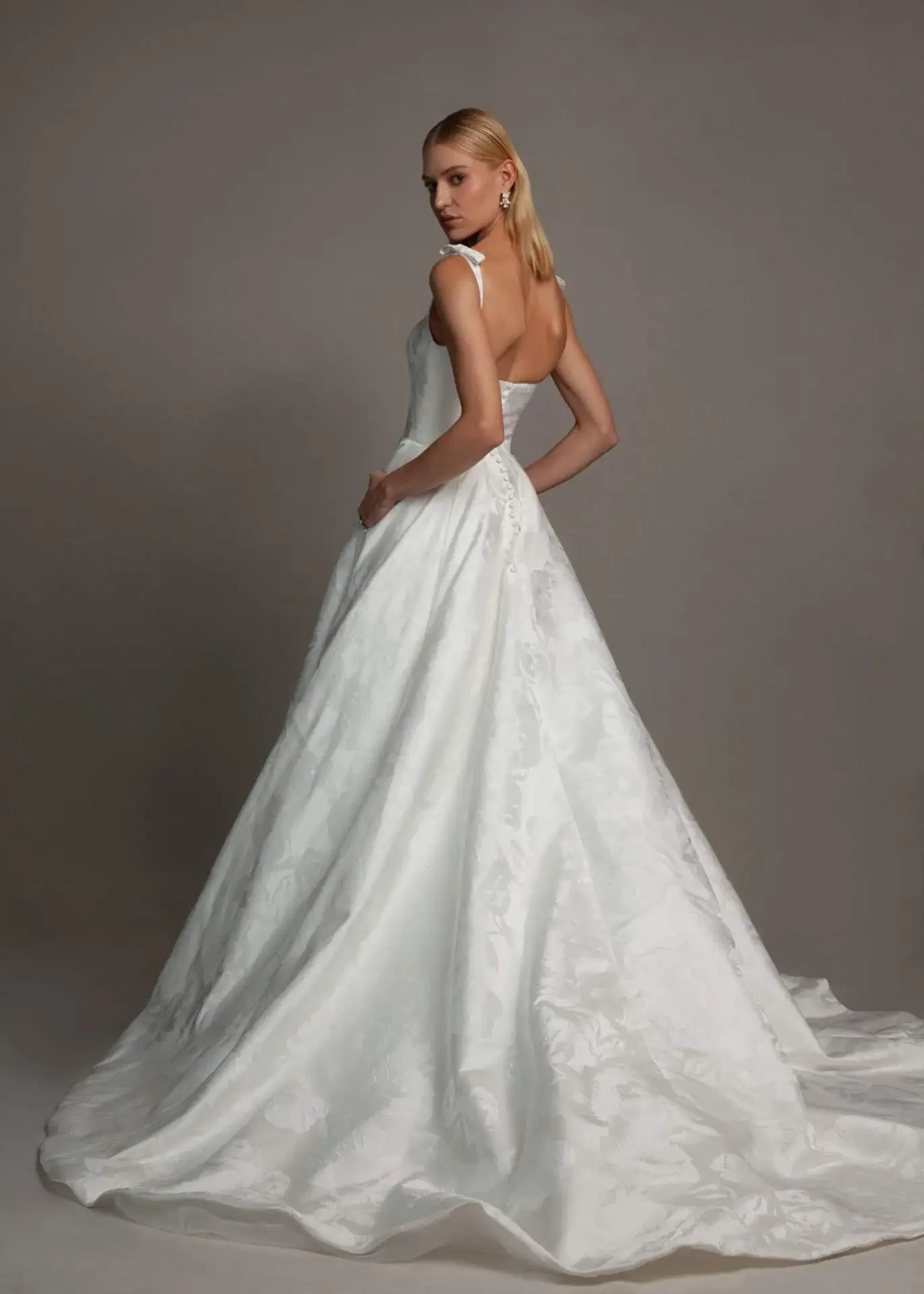 A woman in a white wedding dress with a full skirt and a fitted bodice with shoulder straps, standing against a plain gray background.