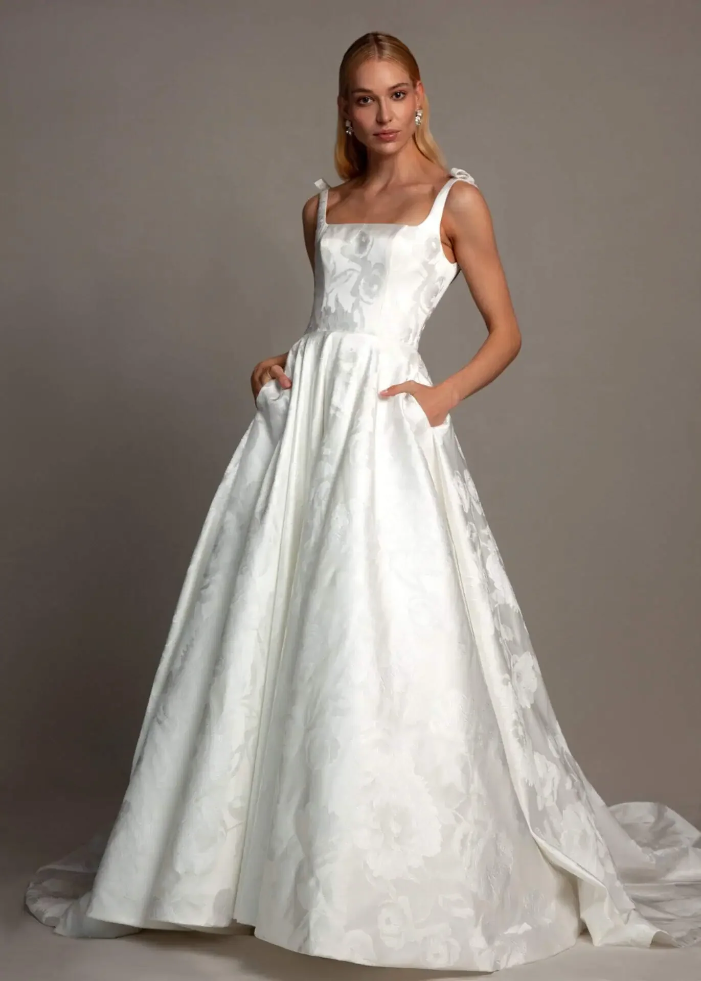 A woman in a sleeveless white wedding gown with a full skirt and pockets, standing against a plain gray background.