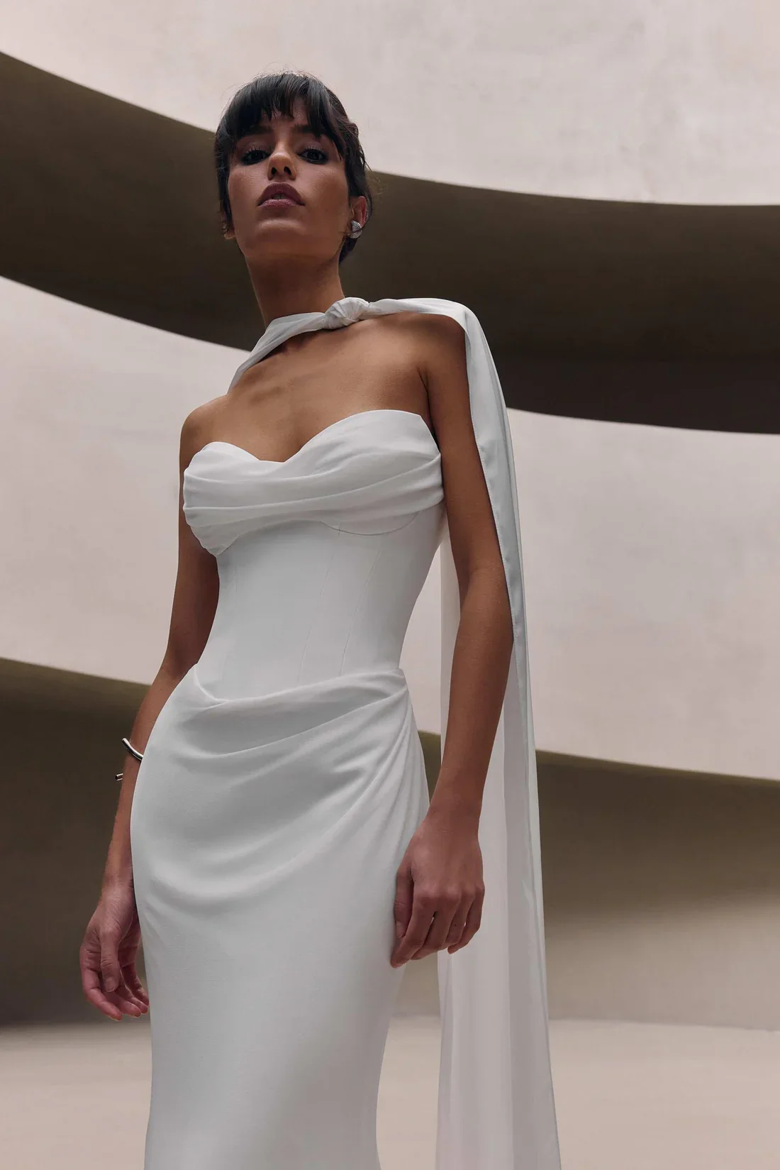 A woman wearing a white, strapless gown with a matching scarf over her shoulder, standing in front of modern, curved architectural background.