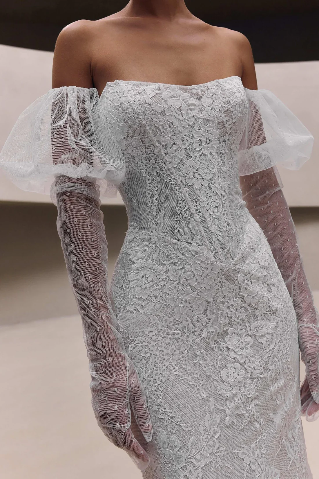 A woman wearing a white, lace wedding dress with puffed tulle sleeves and sheer, dotted, long lace gloves.