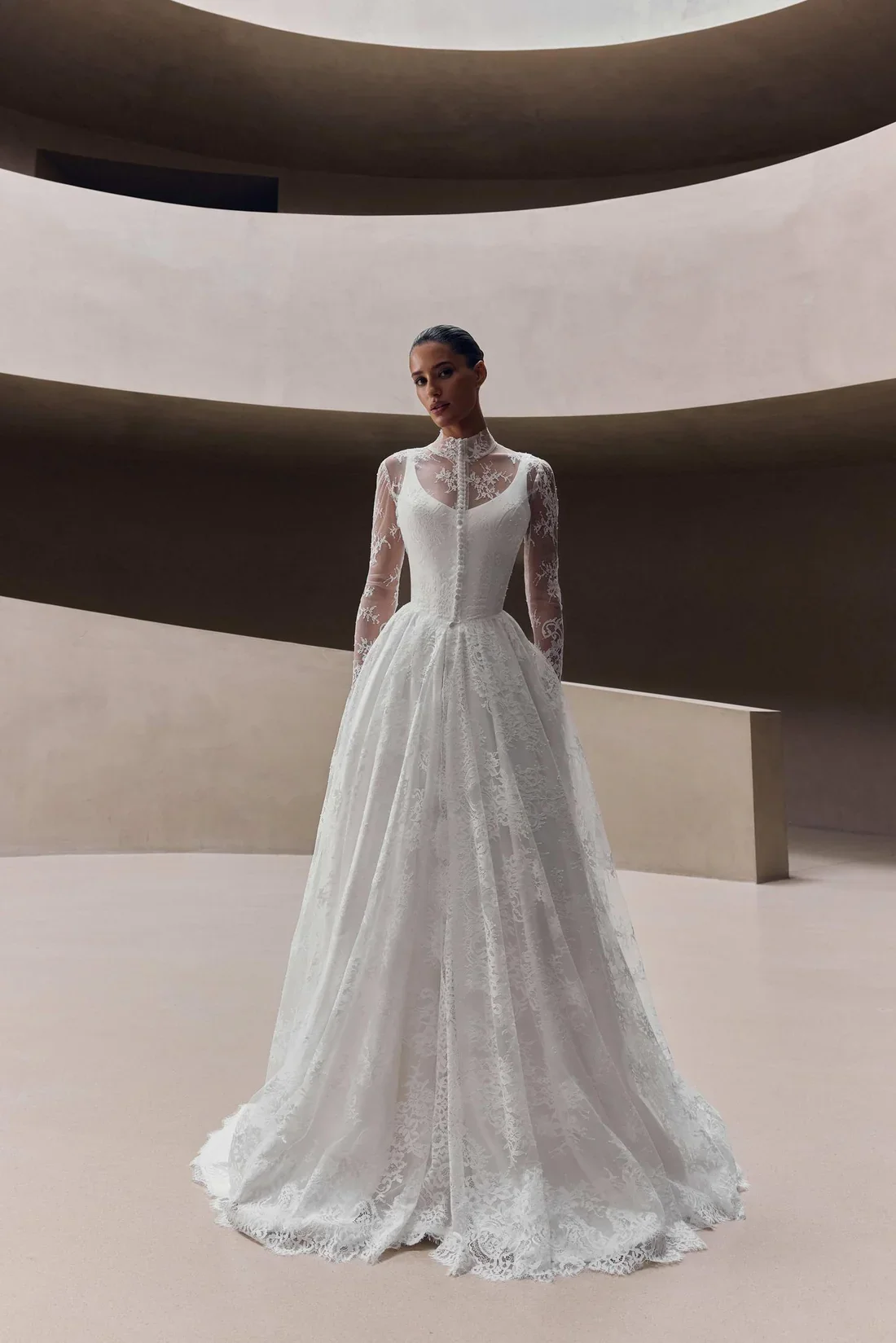 A woman in an elegant white lace wedding dress with long sleeves and a high neckline, standing in a minimalist modern space with curved beige walls.