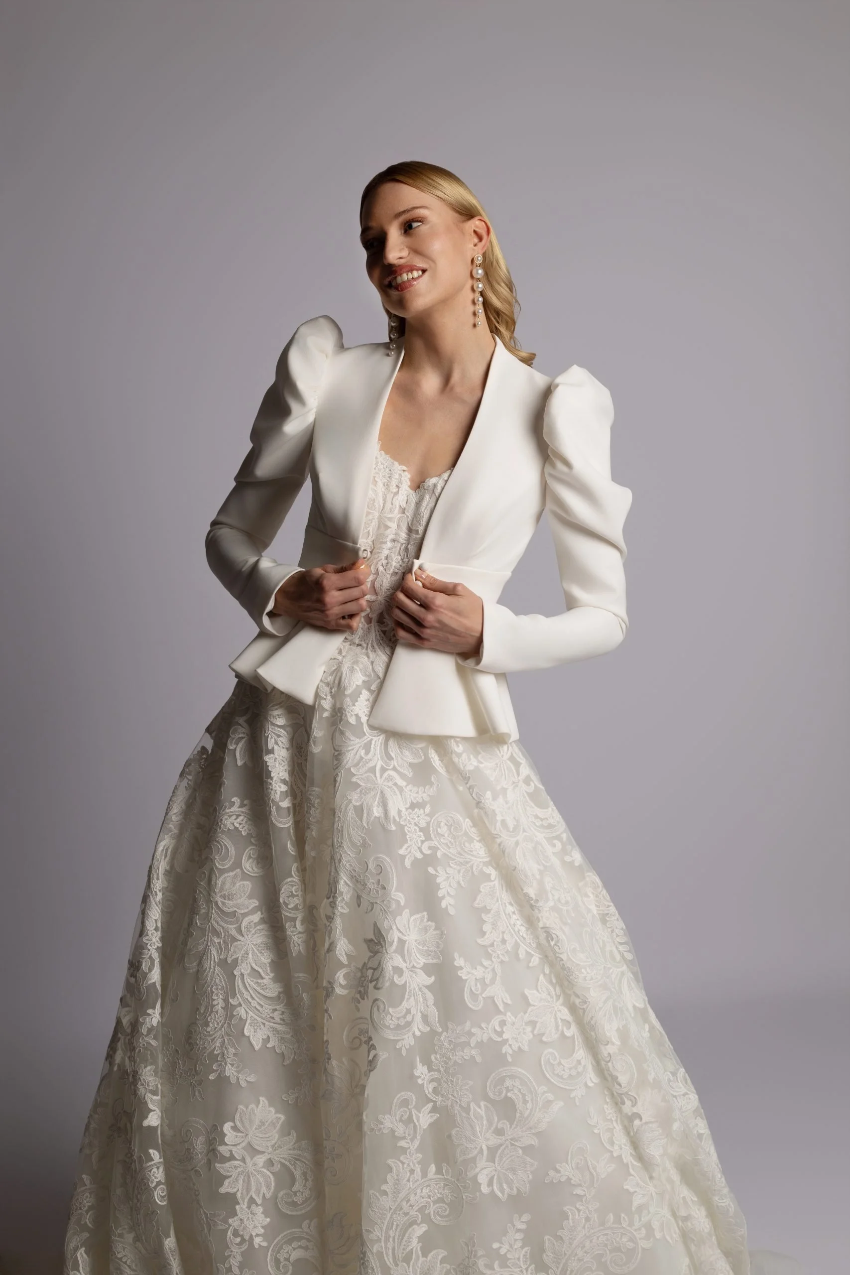 Woman in white wedding dress and blazer posing against a gray background.
