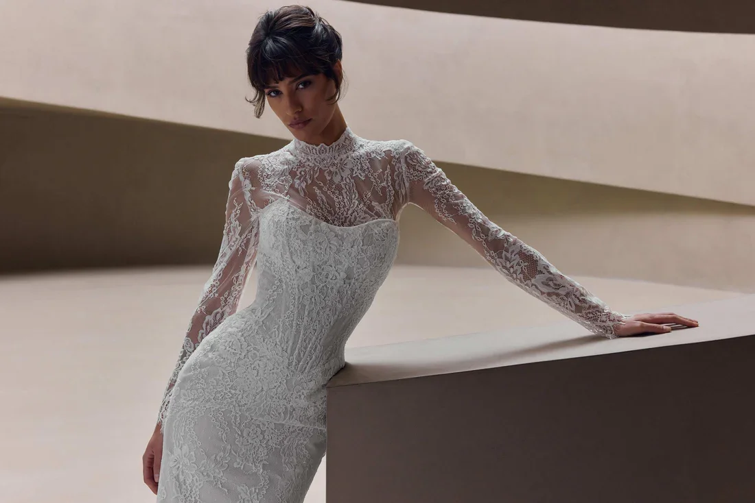 Woman wearing a white lace wedding dress with long sleeves and imprinted lace details, standing in a neutral, minimalistic setting.